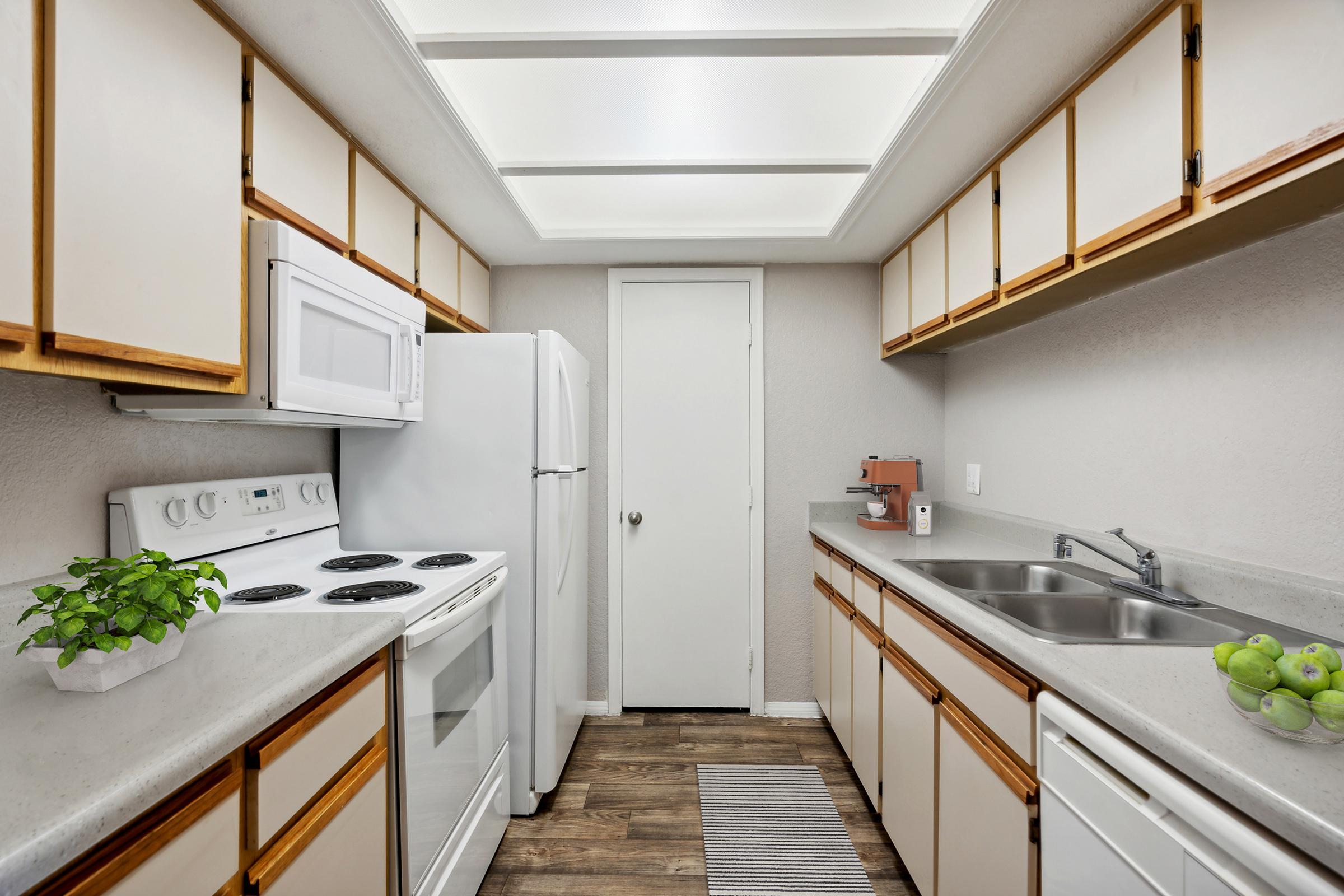 A modern kitchen featuring white cabinetry, a white refrigerator, and an electric stove. The countertops are gray with a sink and double basins. A small green plant decorates the space, and a few green apples are placed near the sink. Natural light filters in through the ceiling, enhancing the bright and clean atmosphere.
