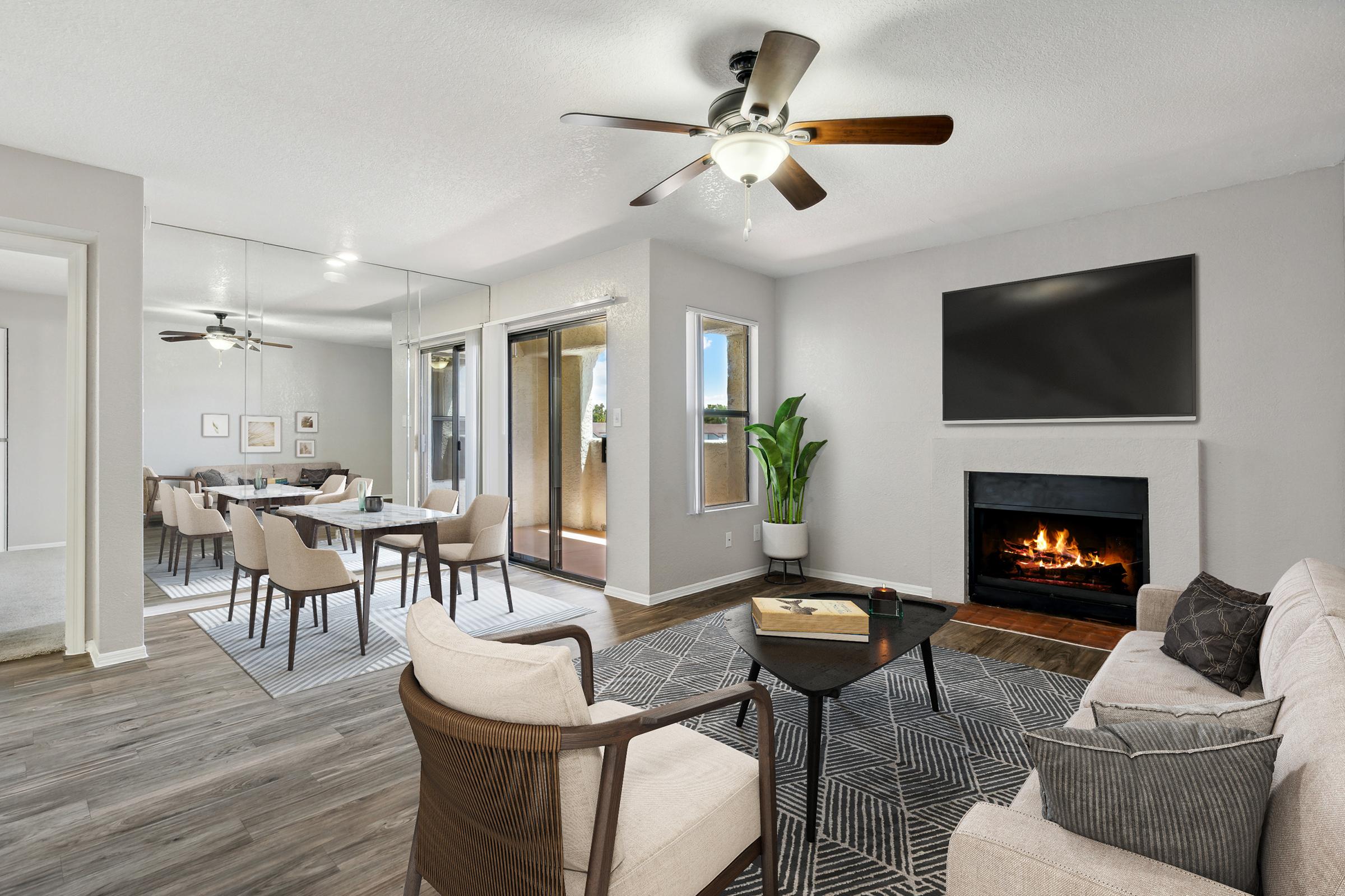 A stylish living room featuring a comfortable beige sofa, a dark wood coffee table, and a cozy fireplace. A large wall-mounted TV hangs above the fireplace. Natural light fills the space, highlighting a patterned rug and a potted plant. A dining area with a modern table and chairs is visible in the background, with large windows showcasing the view.