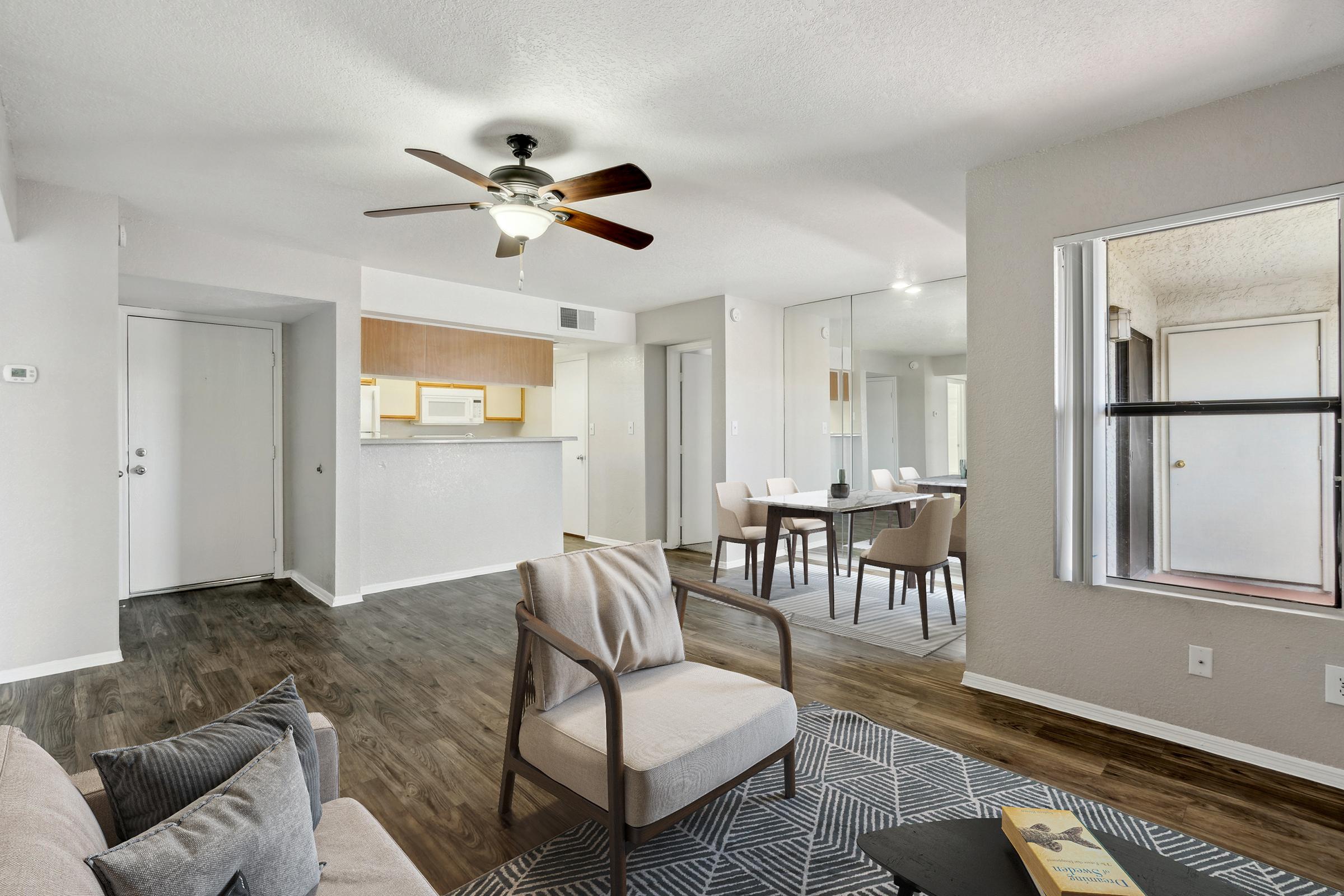 Modern living room with light-colored walls, a ceiling fan, and hardwood flooring. A cozy sofa and armchair sit on a patterned rug, with a dining area in the background featuring a table and chairs. The space includes a kitchen area with a bar counter. Natural light streams in through large windows.
