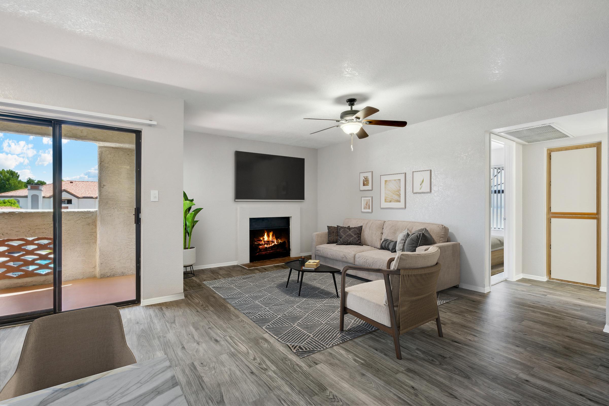 A modern living room featuring a gray sofa with decorative pillows, a sleek coffee table, and a ceiling fan. There's a flat-screen TV mounted on the wall above a stylish fireplace. Large windows allow natural light to fill the space, complemented by a plant and contemporary artwork.