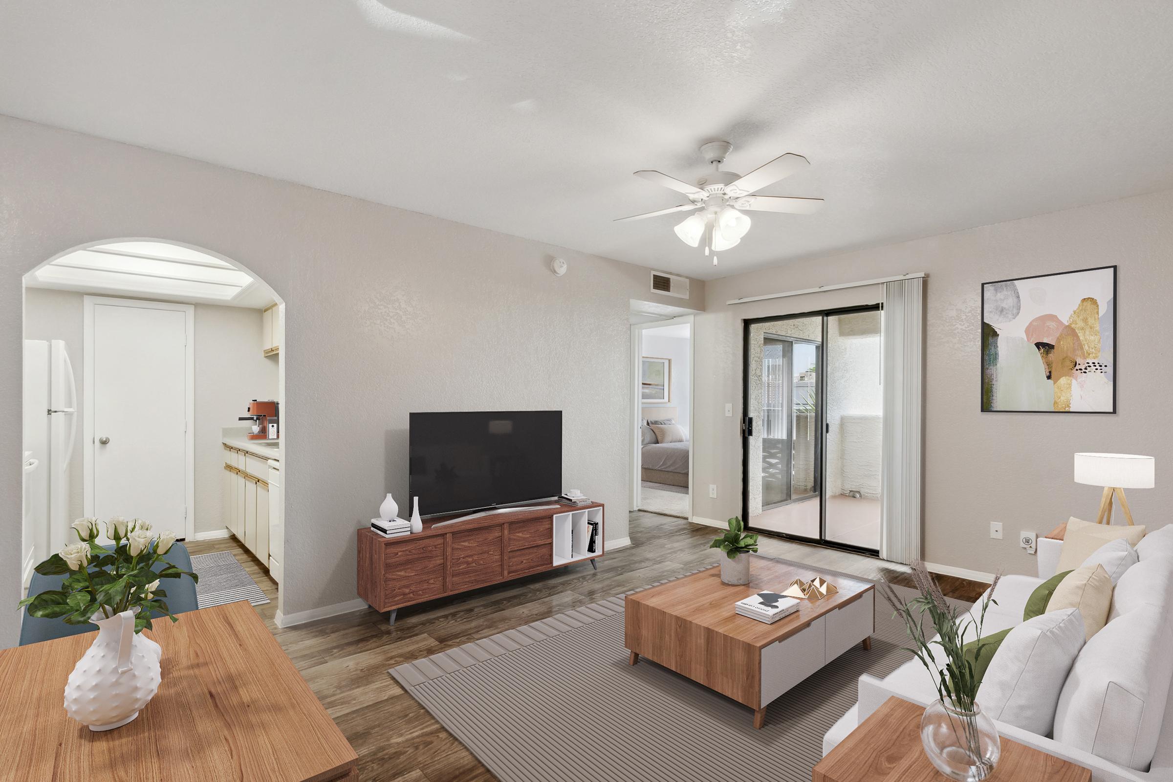 A cozy living room featuring a modern design with a large TV on a wooden stand, a stylish coffee table, and a comfortable sofa. Natural light pours in through sliding glass doors leading to a patio. The room is decorated with simple art and a small vase of flowers, providing a welcoming atmosphere.