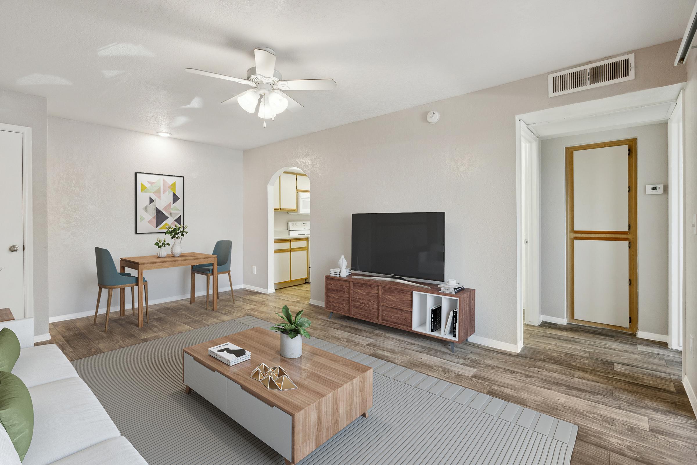 A modern living room featuring a neutral color palette, with a white sofa, a wooden coffee table, and a large TV on a console. In the background, there's a small dining area with a table and chairs, an arched doorway leading to a kitchen, and a door to another room. The space is brightly lit with a ceiling fan overhead.