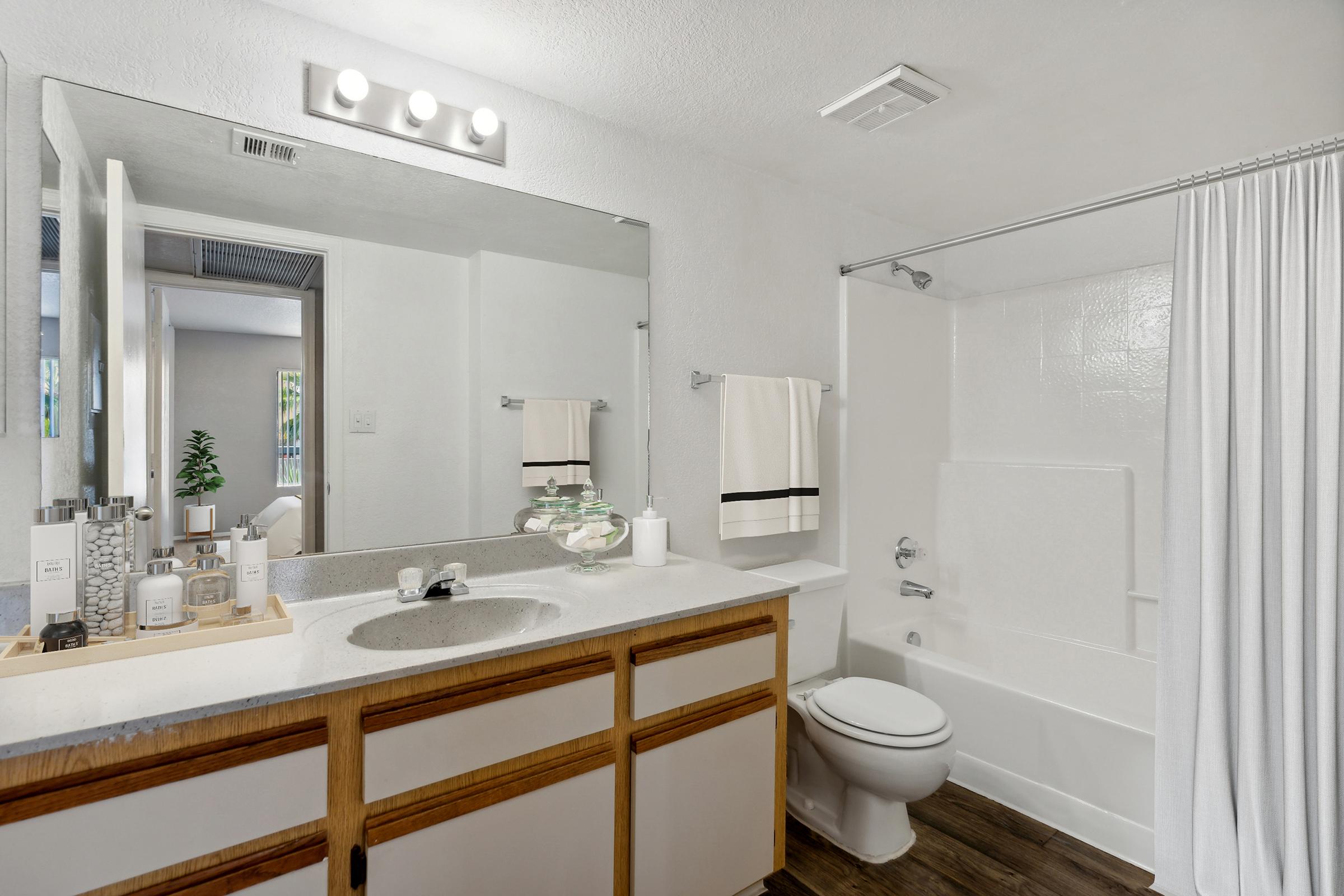 A clean and modern bathroom featuring a white bathtub and shower, a large mirror above a sink with a wooden cabinet, neatly arranged toiletries, towels hanging on a rack, and natural light coming in from a nearby window. The walls and floor are in neutral tones, creating a bright and spacious feel.