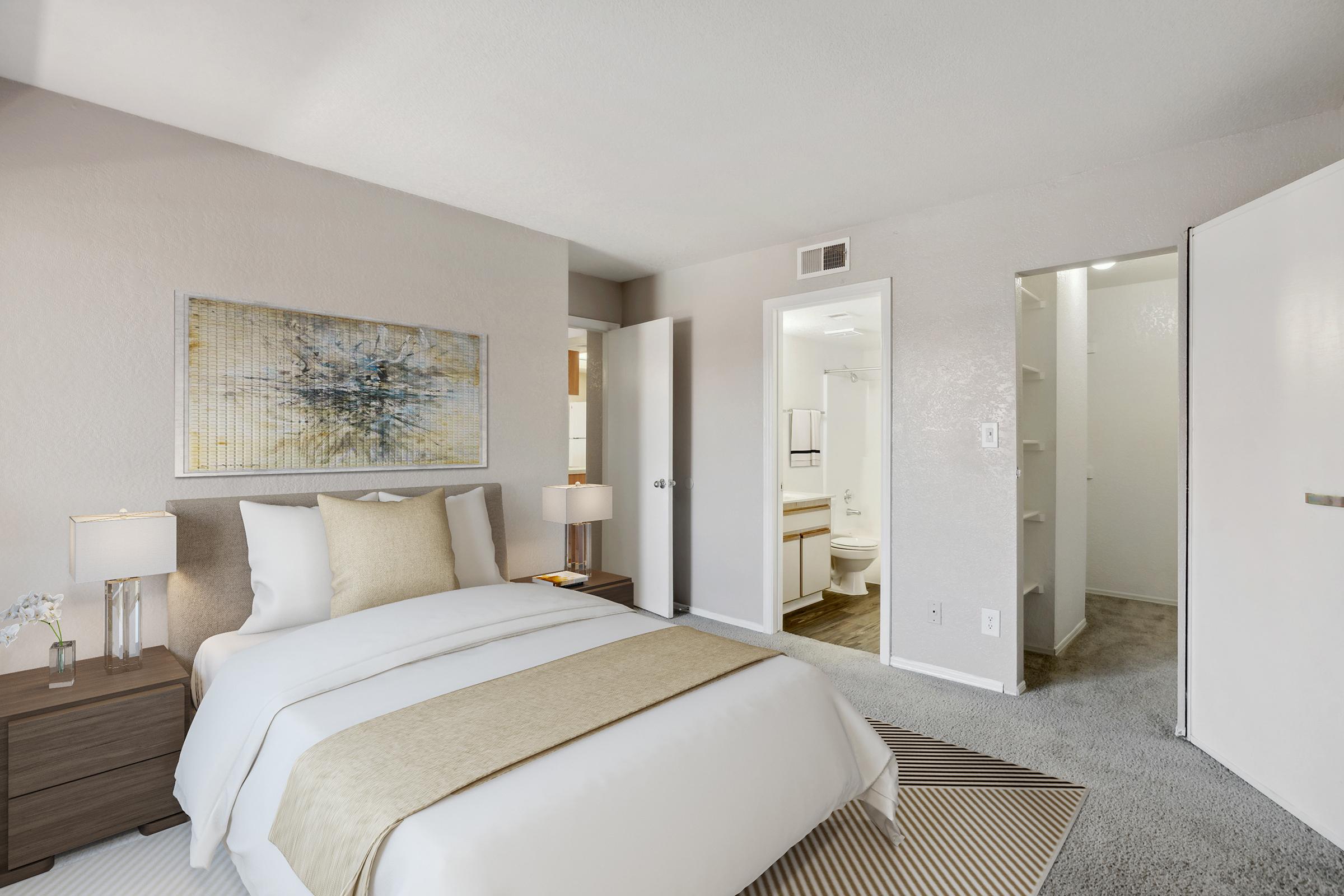 A cozy and modern bedroom featuring a neatly made bed with cream-colored bedding, a light gray carpet, and a bedside table with a decorative lamp. A bathroom is visible in the background, showing a white door and light fixtures. The walls are painted in soft neutral tones, enhancing the room's tranquility.