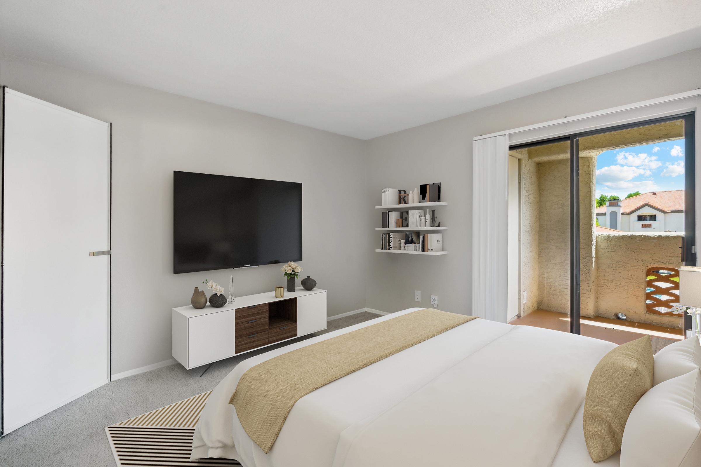 A modern bedroom featuring a queen-sized bed with a light-colored comforter, a sleek white nightstand, and a wall-mounted flat-screen TV. A bookshelf mounted on the wall holds decorative items. A sliding glass door opens to a small balcony with bright outdoor scenery. Neutral tones create a calming atmosphere.