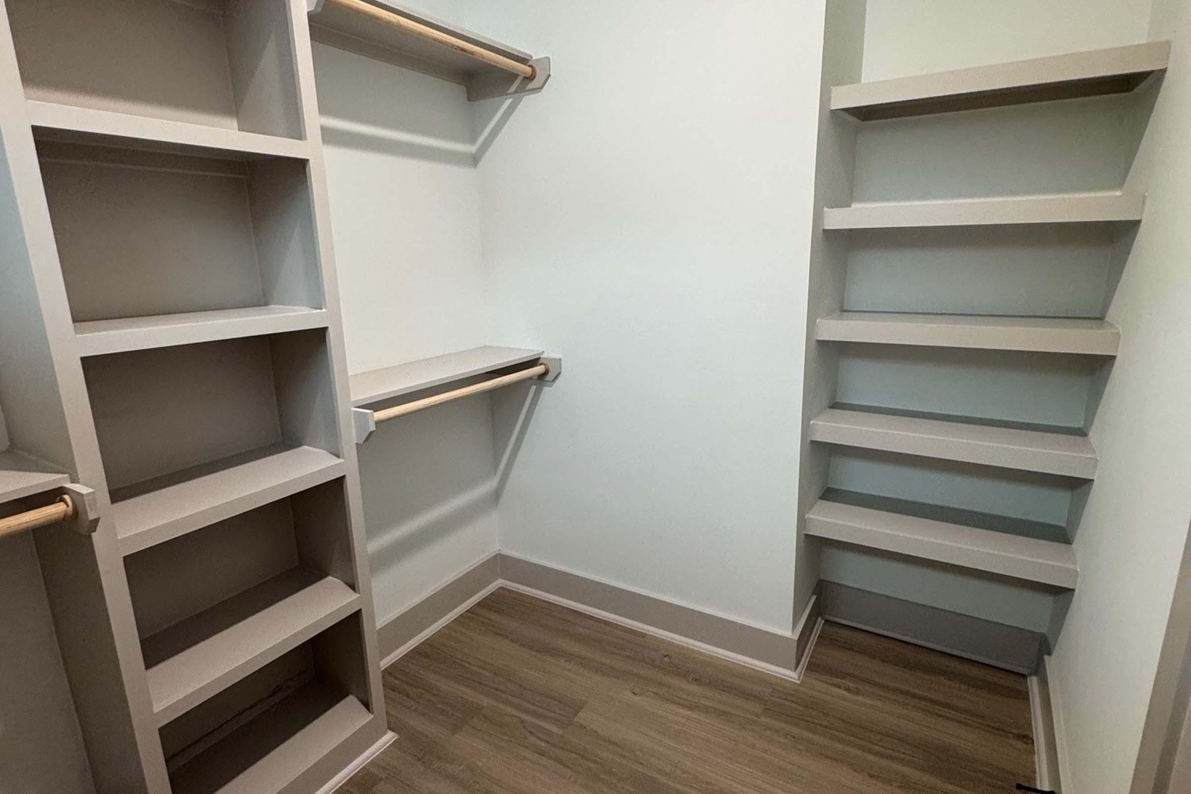A well-organized empty closet featuring light gray shelving units and a hanging rod. The walls and flooring are neutral tones, creating a bright and spacious appearance, ideal for storage or customization.