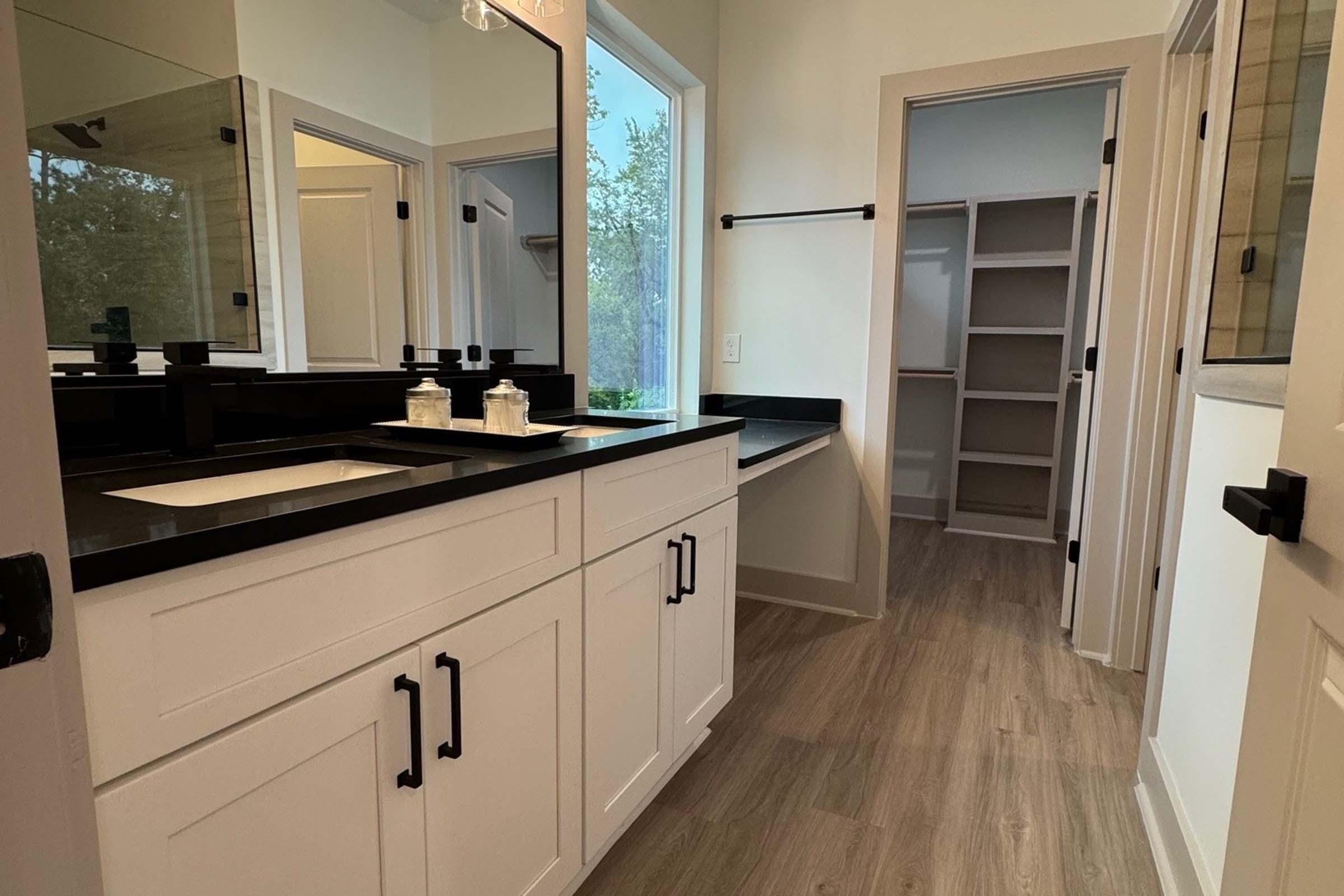 Spacious modern bathroom featuring a black countertop with two sinks, light-colored cabinetry, and ample storage space. Large mirrors reflect natural light from window, and a walk-in closet is visible through an open door. Stylish fixtures and a clean, minimalist design enhance the aesthetic.