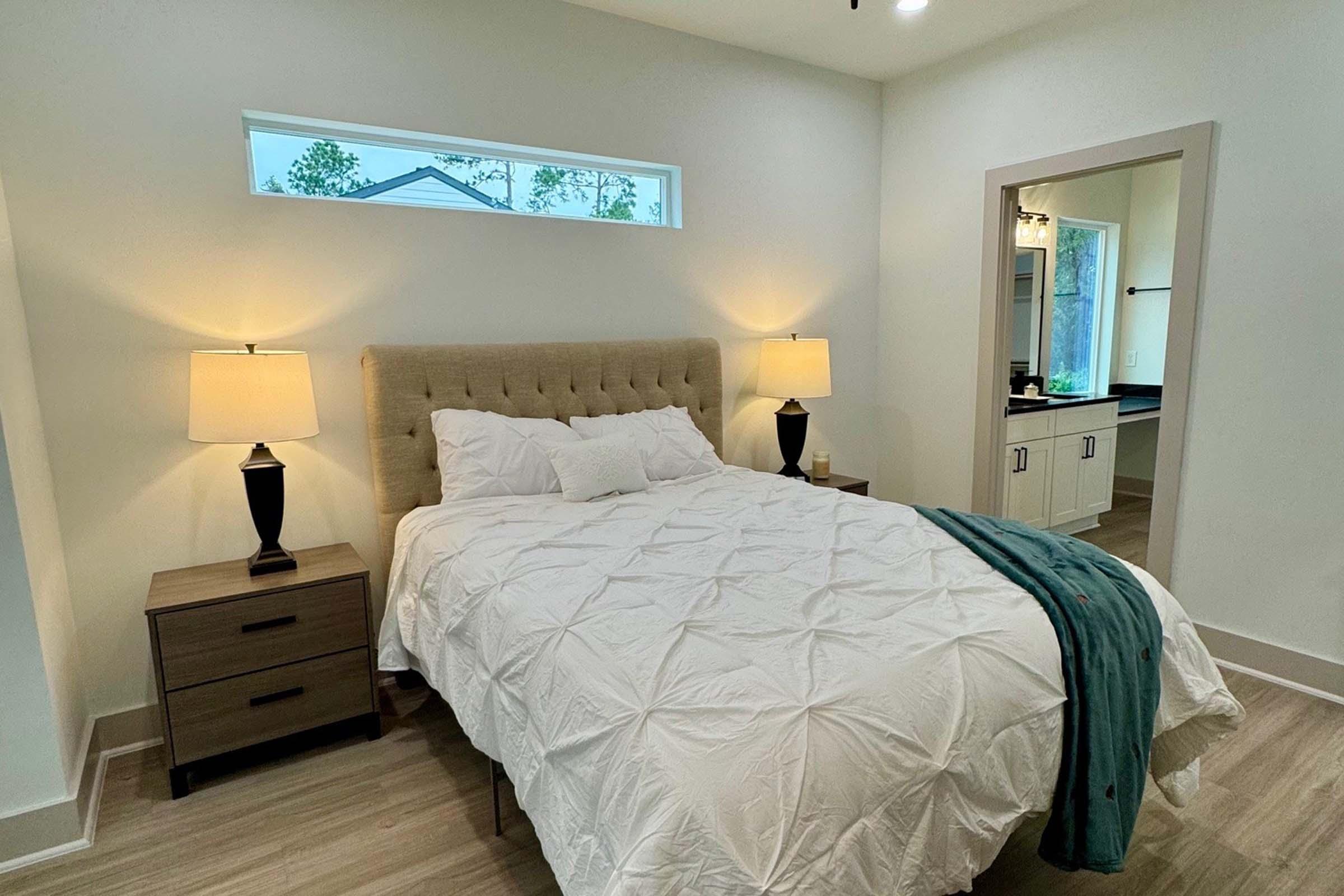 A cozy bedroom featuring a queen-sized bed with a light-colored tufted headboard and a white quilted bedspread. Two bedside lamps with black bases are on either side. A mirrored closet door reflects a bright, airy space with a window allowing natural light to flow in.