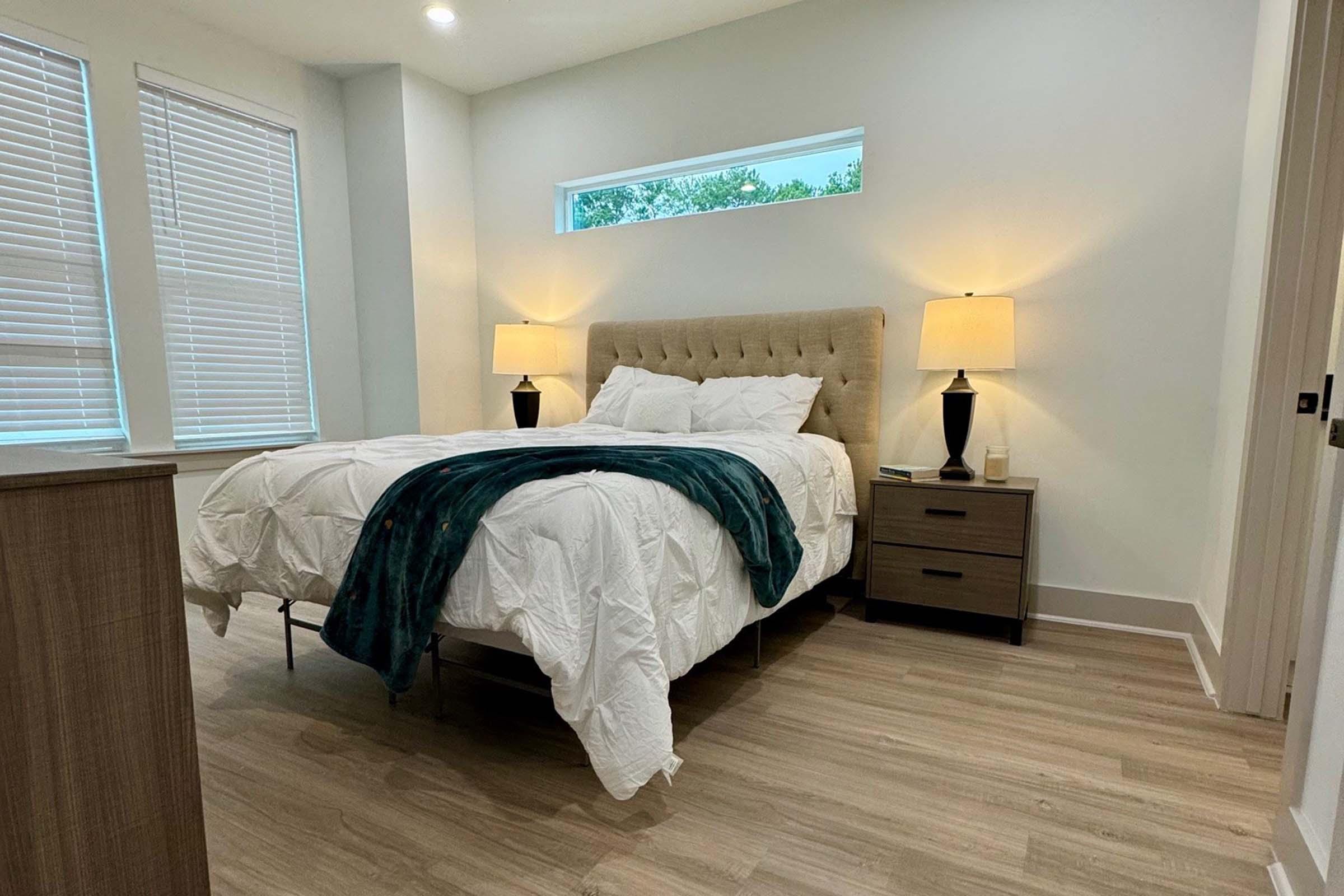 A modern bedroom featuring a plush, light-colored bed with white bedding and a teal throw blanket. There are two nightstands with black lamps on either side, and large windows with blinds providing natural light. The flooring is wooden, and the walls are painted a soft, neutral color.