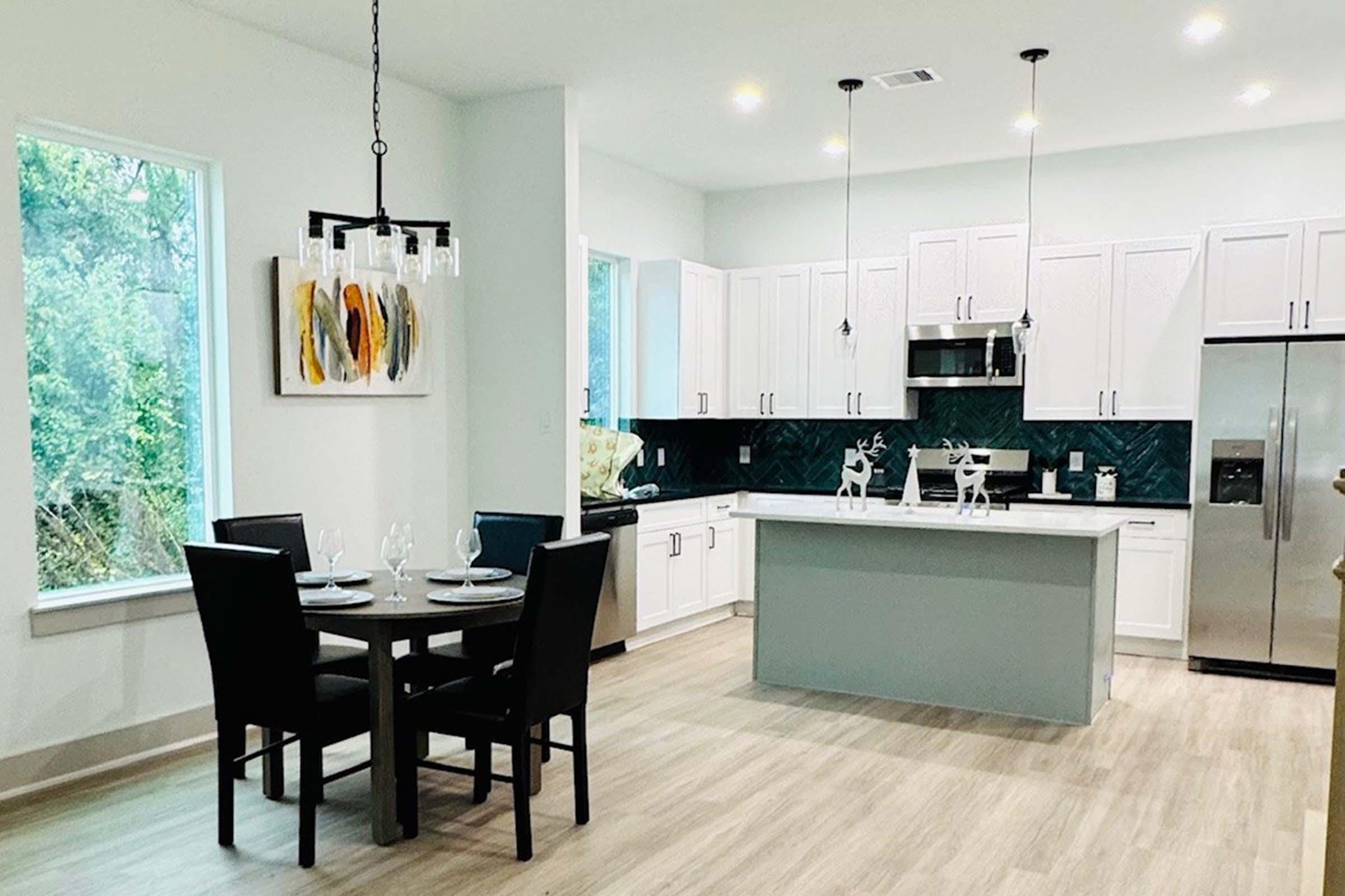 Modern kitchen featuring white cabinets, a teal kitchen island, and silver appliances. A dining table with four black chairs is set for a meal, with plates and glasses. Large windows allow natural light to fill the space, complemented by a contemporary chandelier and abstract artwork on the wall.