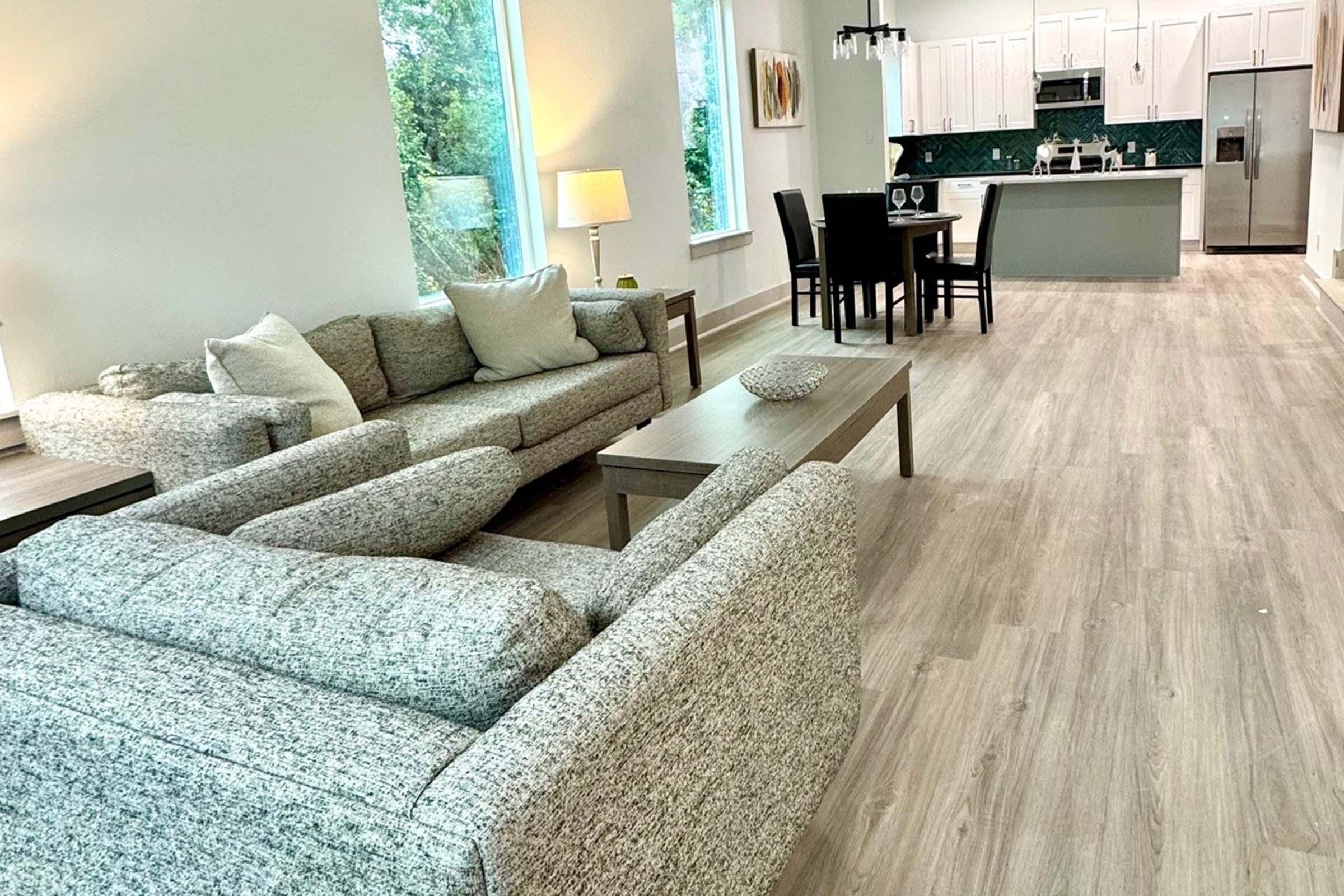 A modern living area featuring a comfortable gray sofa, a coffee table, and a dining table with chairs. Large windows allow natural light to fill the space, highlighting the light wooden flooring and contemporary kitchen in the background. The decor is minimalist and inviting.