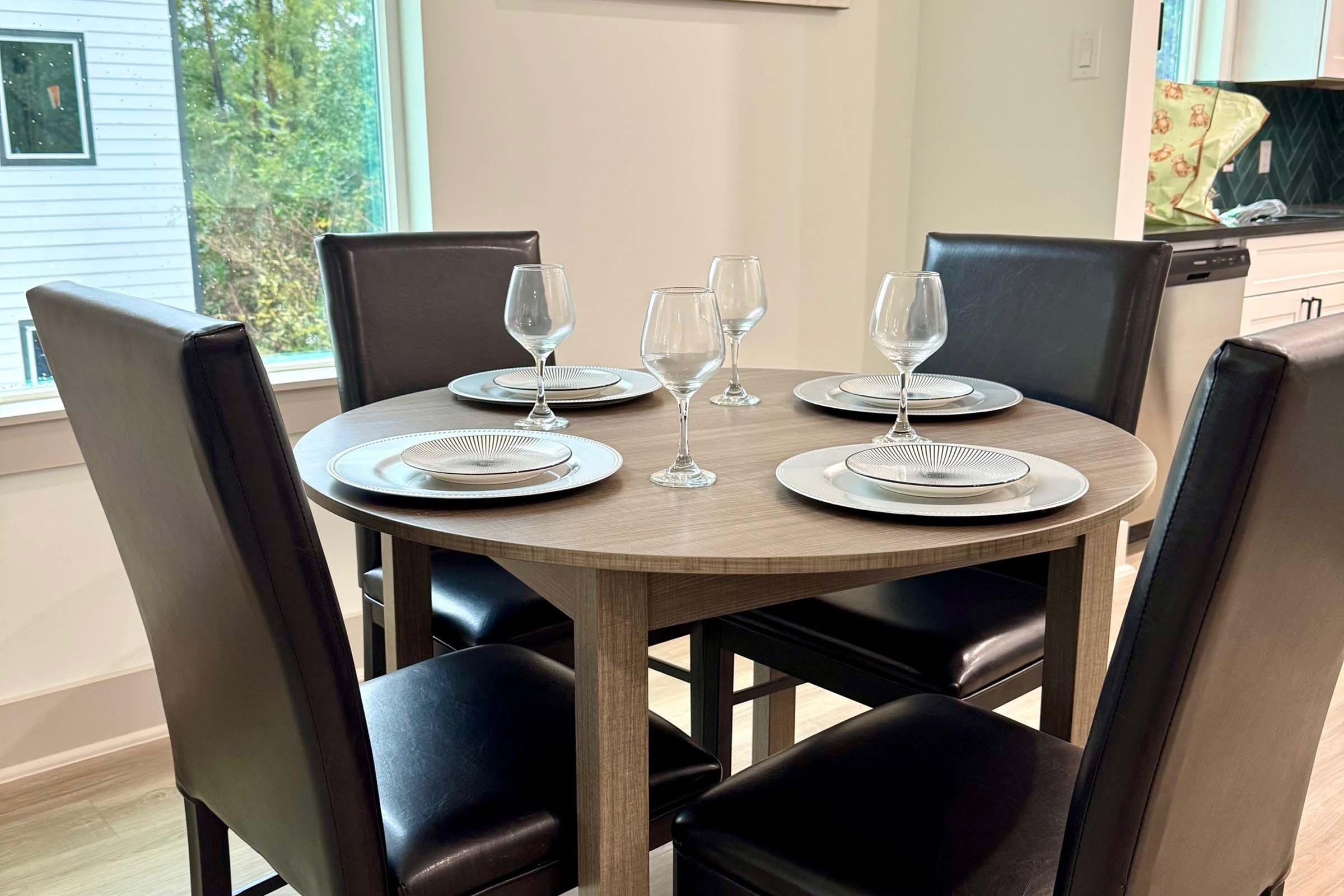 A round dining table set for four, featuring elegant plates and wine glasses. The table is surrounded by black leather chairs, and there is a bright, airy atmosphere with natural light coming through a nearby window. The background shows a modern kitchen design.