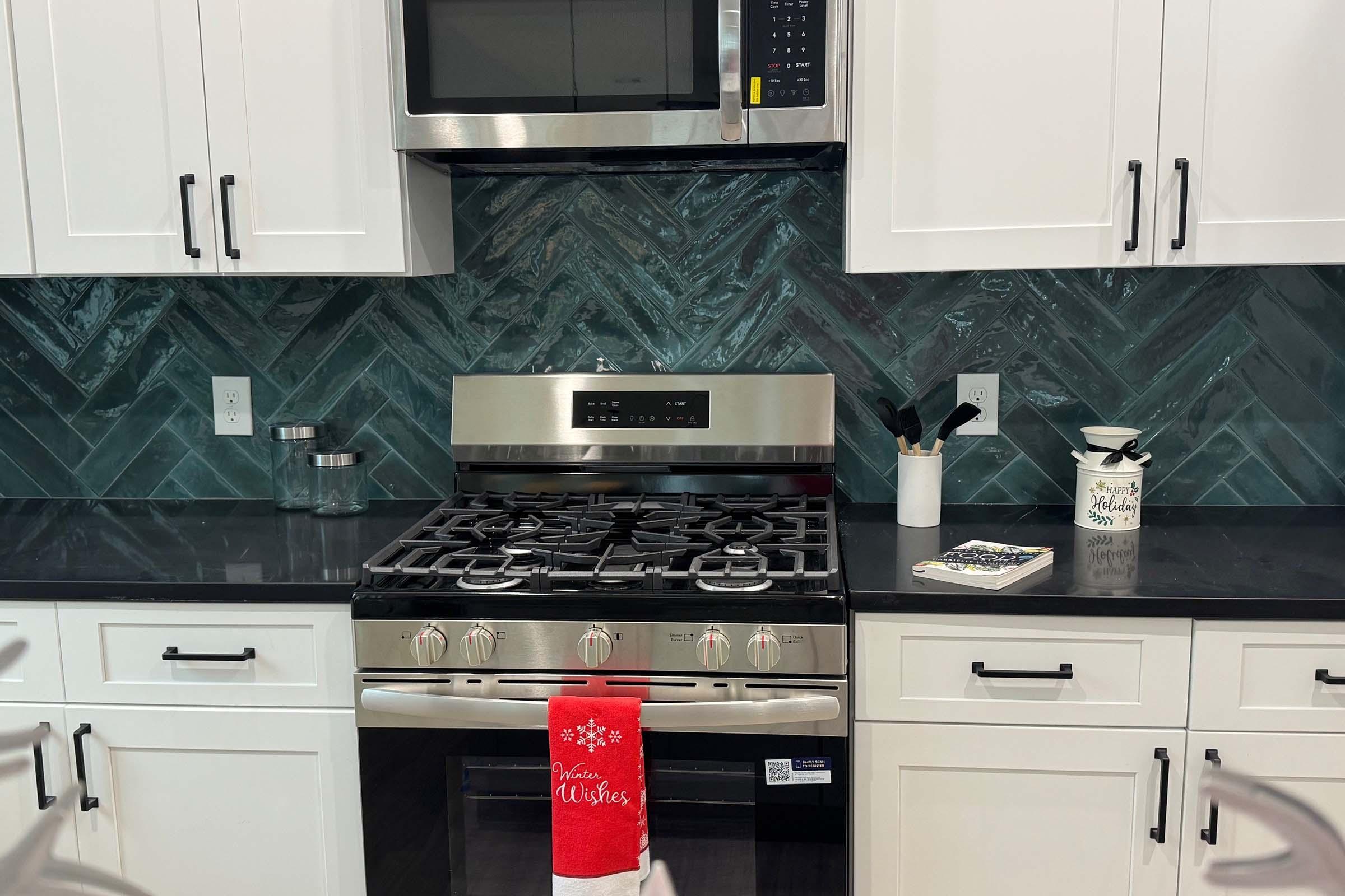 A modern kitchen featuring a stainless steel stove and microwave above it. The backsplash is made of dark green, herringbone-patterned tile. A red towel with "Christmas Wishes" is hanging from the stove. White cabinets and a black countertop complete the look, with utensils and a decorative jar on the counter.