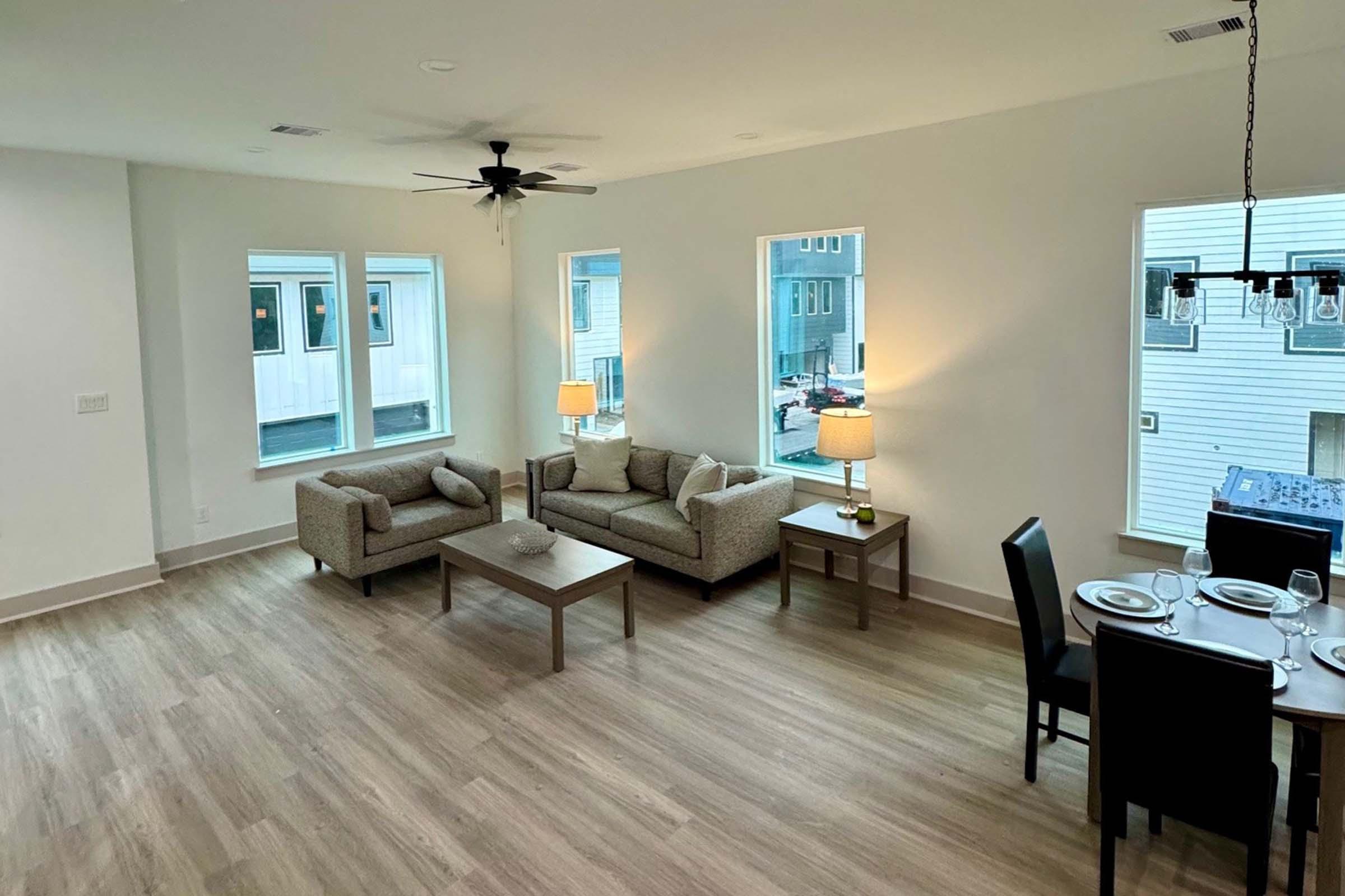 A modern, well-lit living room featuring two gray sofas, a coffee table, and two lamps. Large windows provide natural light, and a dining area with a black table and chairs is visible in the background. The flooring is light wood, and the walls are painted white, creating a spacious feel.