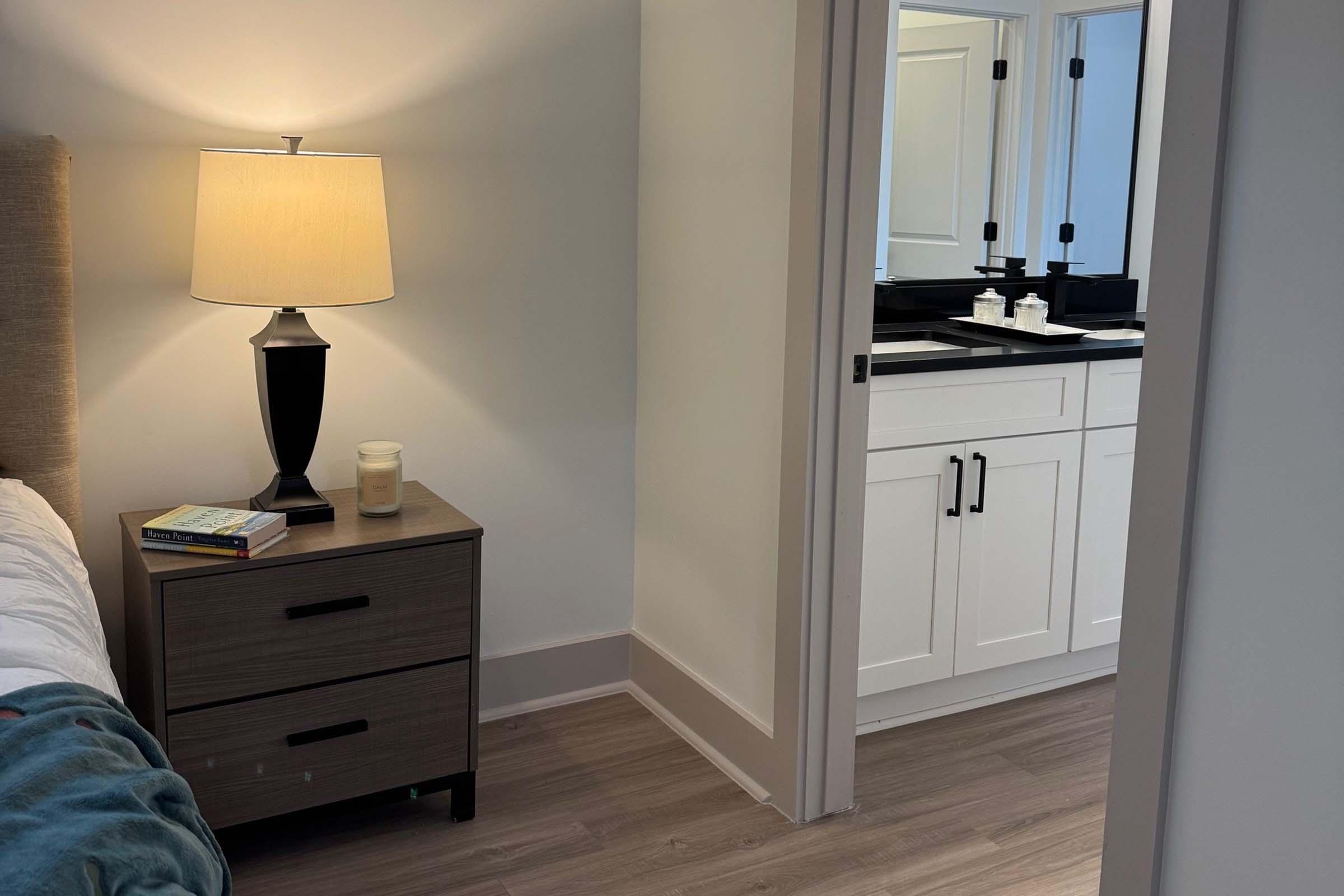 A well-lit bedroom featuring a bedside table with a lamp, books, and a candle. In the background, a doorway leads to a bathroom with a modern design, including a double vanity and mirrors. The room has light wood flooring and neutral-colored walls. A cozy blanket is casually draped over the bed.