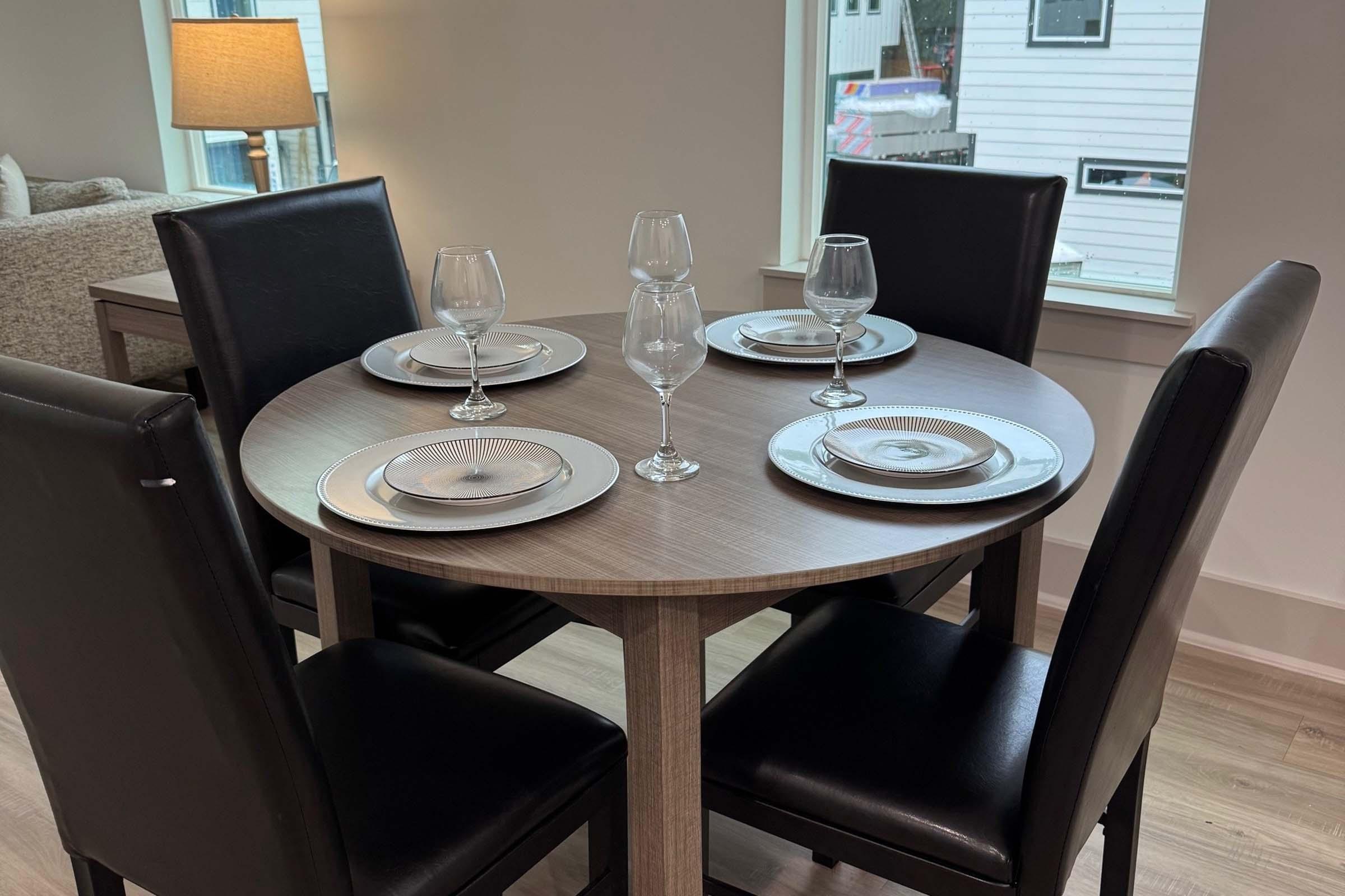 A small round dining table set for four, featuring silver plates, wine glasses, and black padded chairs. The table is positioned near a window with natural light streaming in, and the background shows a cozy living area.