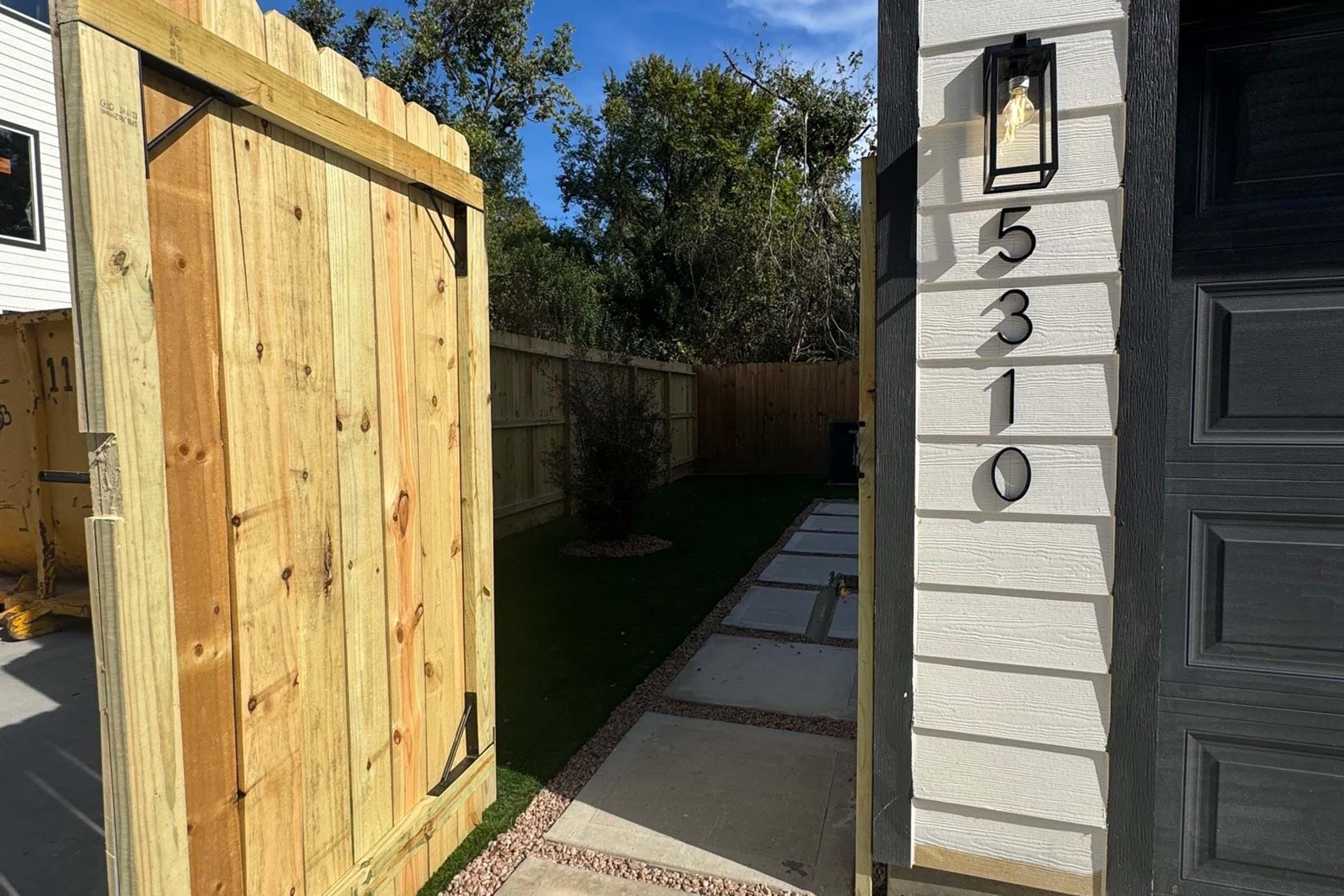 A wooden gate opens to a neatly landscaped path leading to a house with the number "5310" displayed on the wall. The path features stone pavers and is bordered by grass. A small shrub is visible along the side, with trees and blue sky in the background.