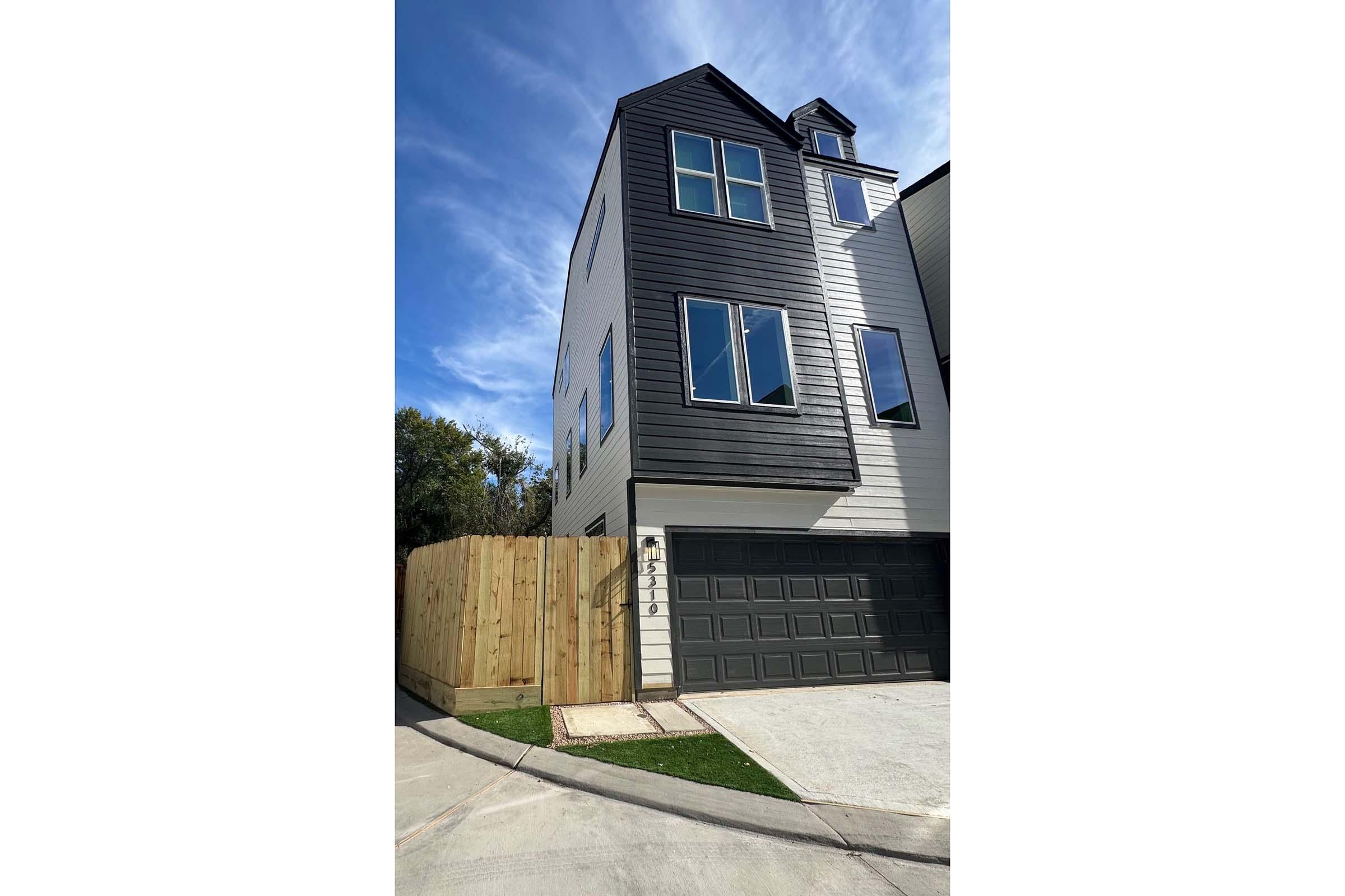 Three-story modern house with a mix of black and white siding, large windows, and a garage. The front yard is enclosed by a wooden fence. The sky is bright blue with a few clouds, and the driveway is paved.