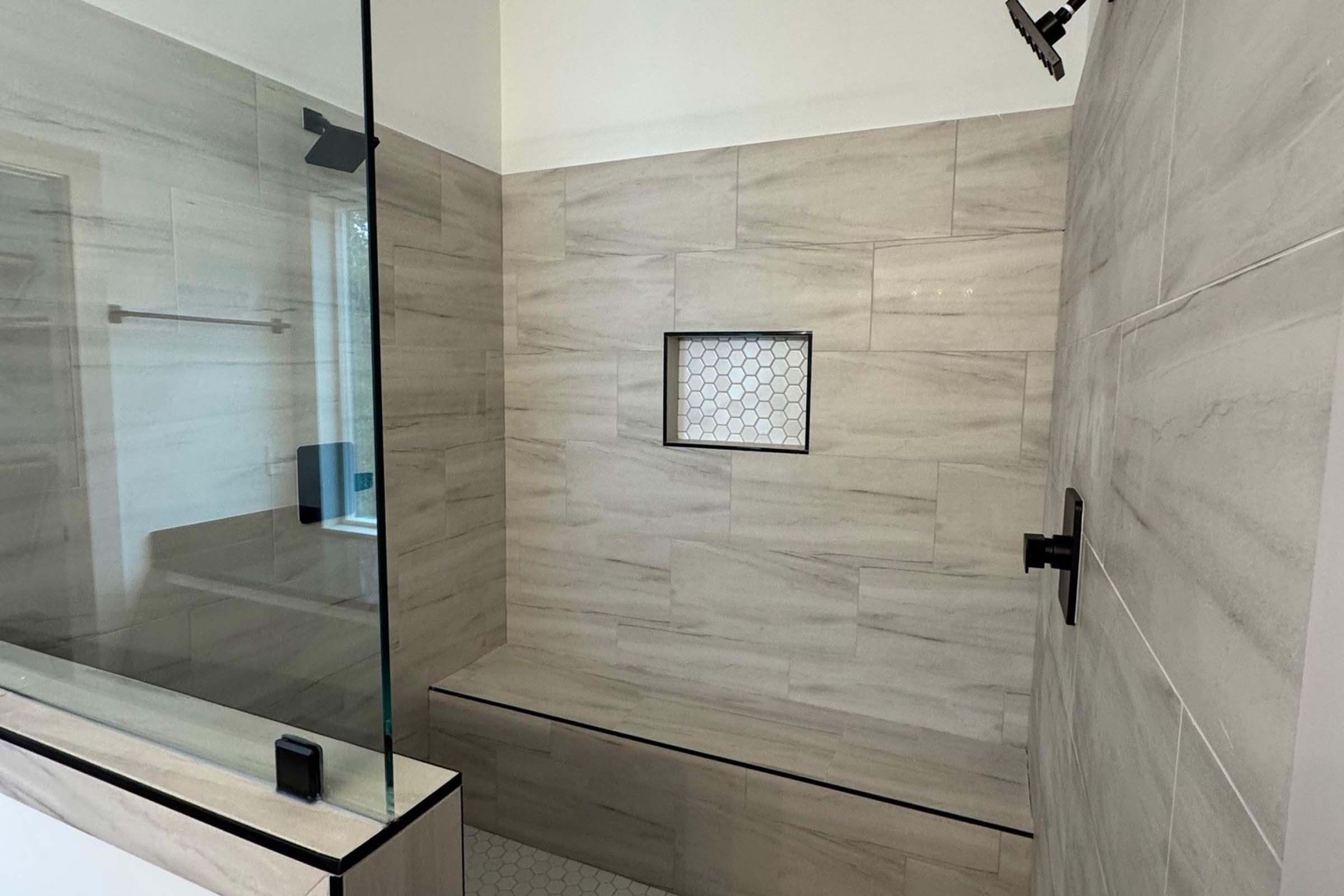 A modern shower featuring glass walls, light-colored stone tiles, and a built-in bench. The shower has a minimalist design with neutral tones and a small niche for toiletries. The floor is tiled with hexagonal patterns, enhancing the contemporary look.