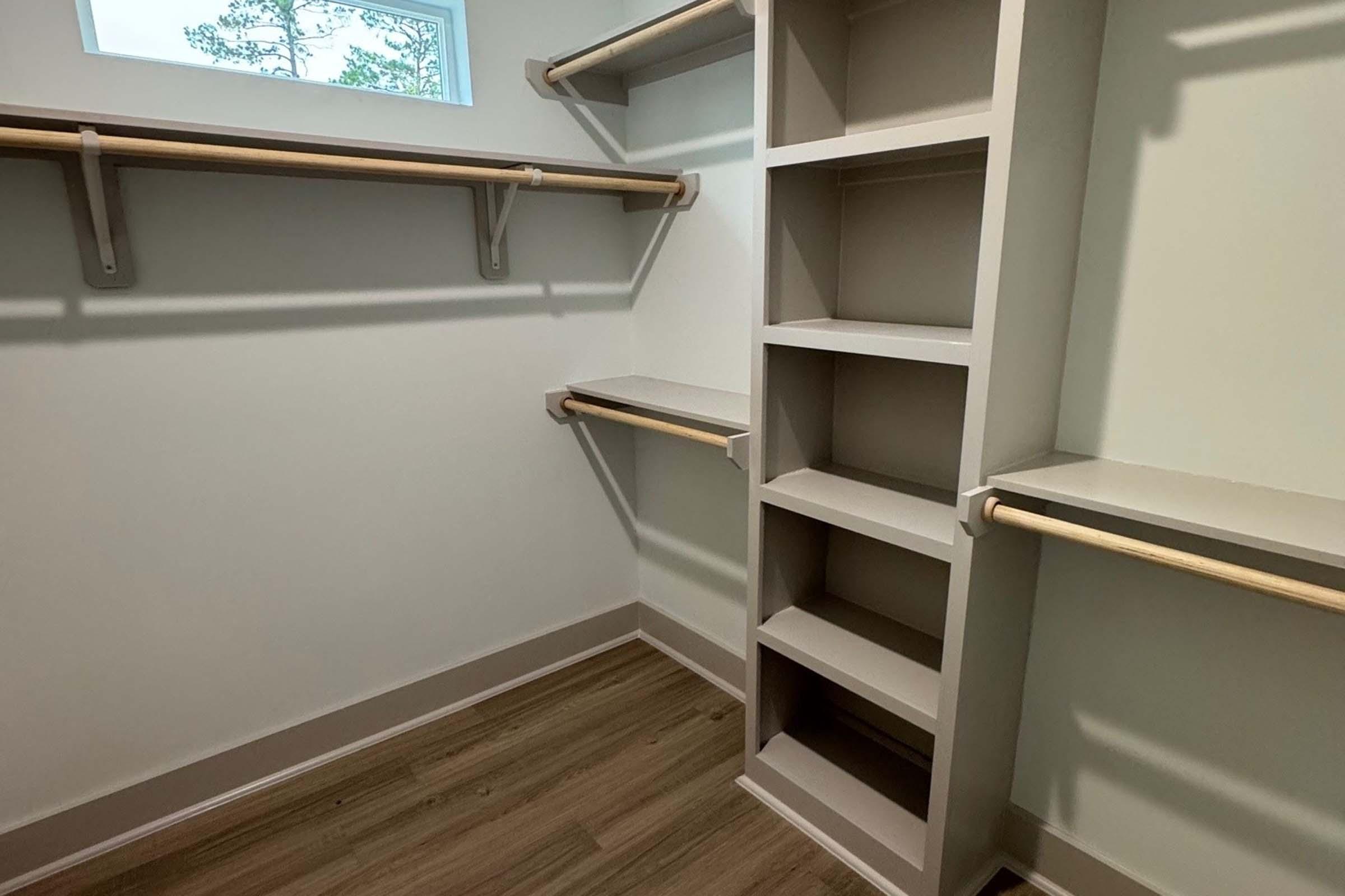A minimalist walk-in closet featuring light-colored walls, wooden flooring, and several hanging rods on the walls. There's a built-in shelving unit on one side, providing storage space, and a small window allowing natural light to illuminate the area.
