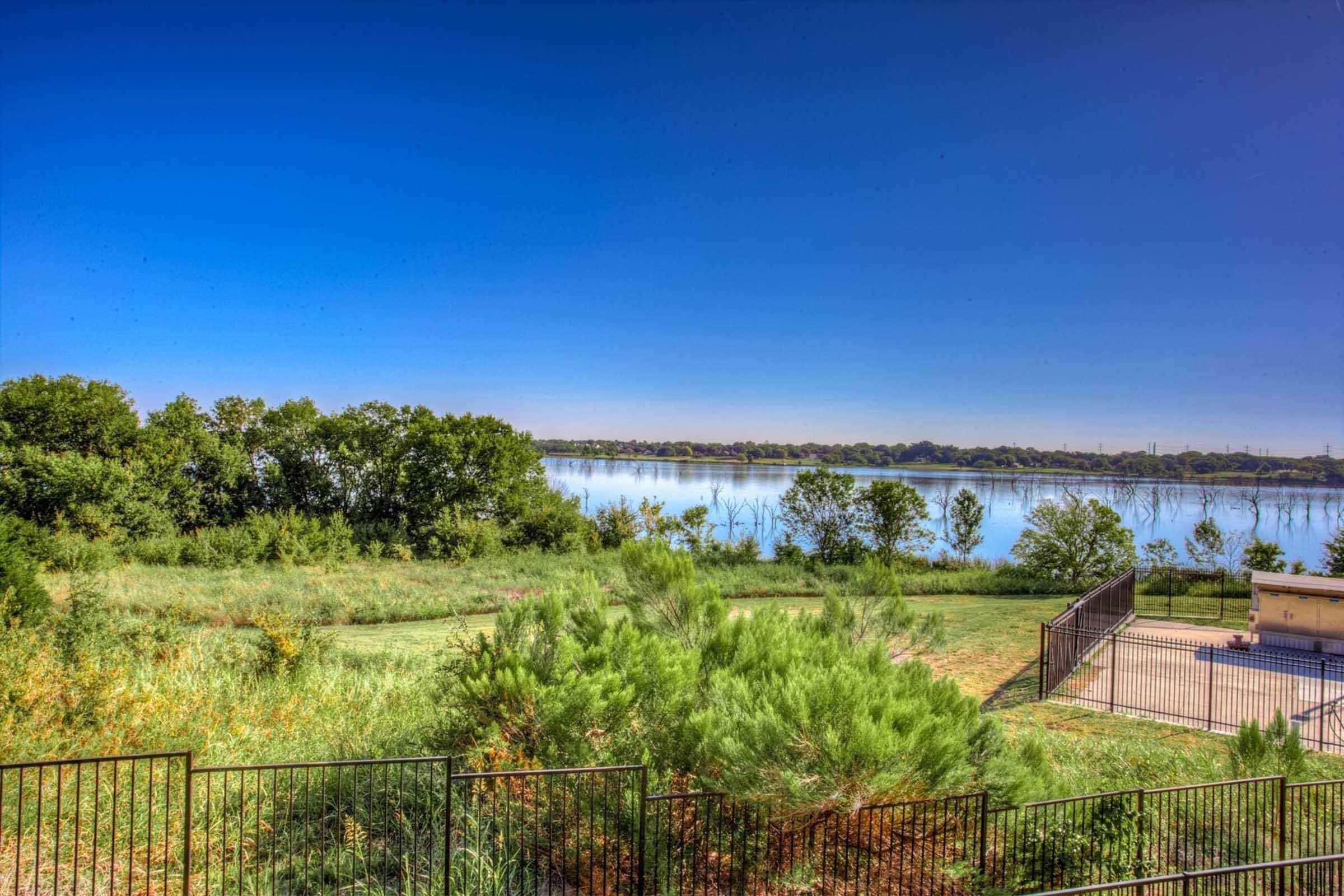 A tranquil view of a calm lake surrounded by lush greenery and trees under a clear blue sky. The foreground features tall grasses and plants, while the distant shoreline reflects the serene waters of the lake. A fenced area is visible on the right side, adding to the peaceful landscape.