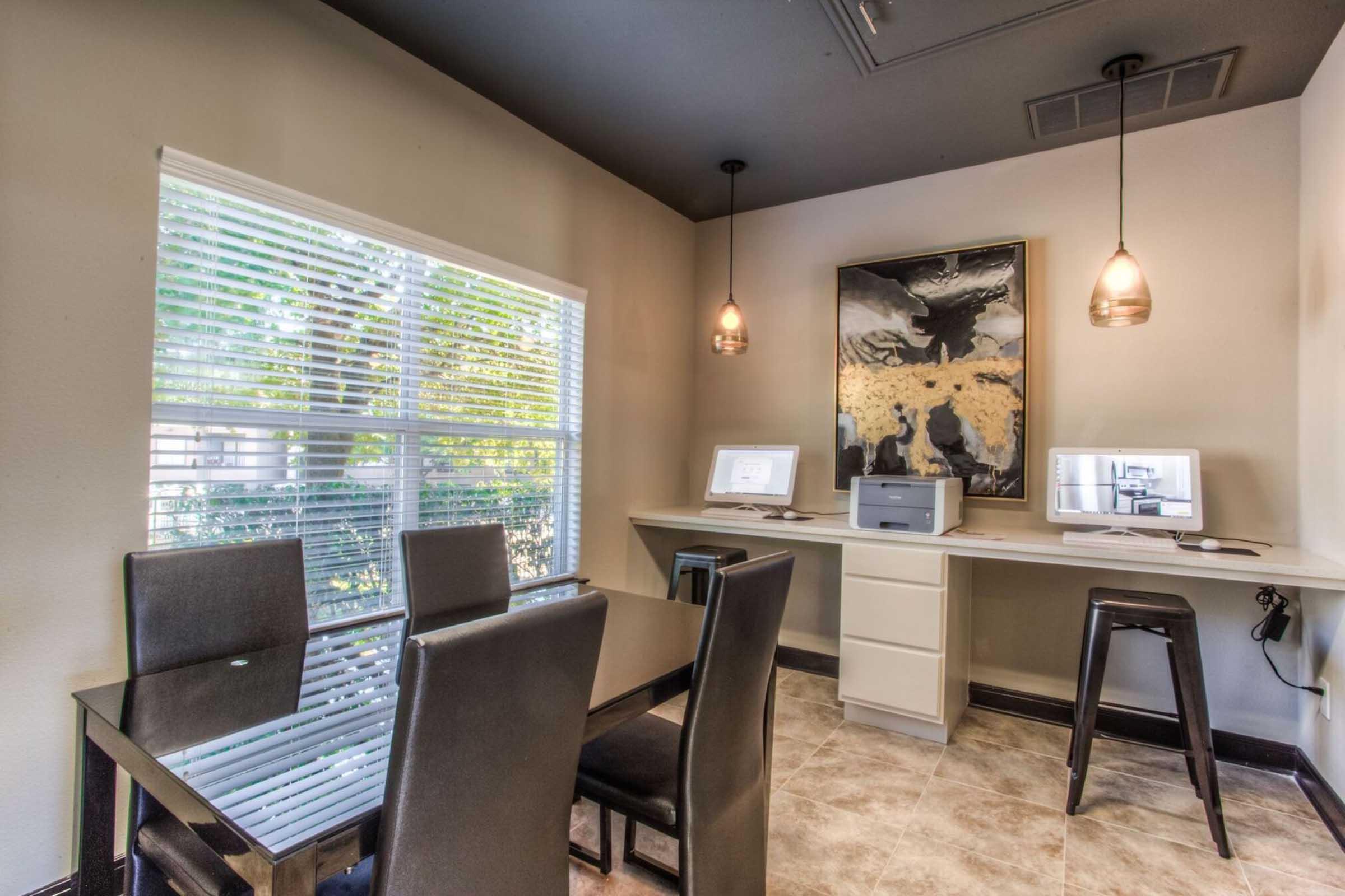 A modern office space featuring a glass-top dining table with six black chairs, a workstation with two computers, a printer, and a large abstract art piece on the wall. Natural light floods in through a window with blinds, enhancing the contemporary decor. The floor is tiled in a neutral color.