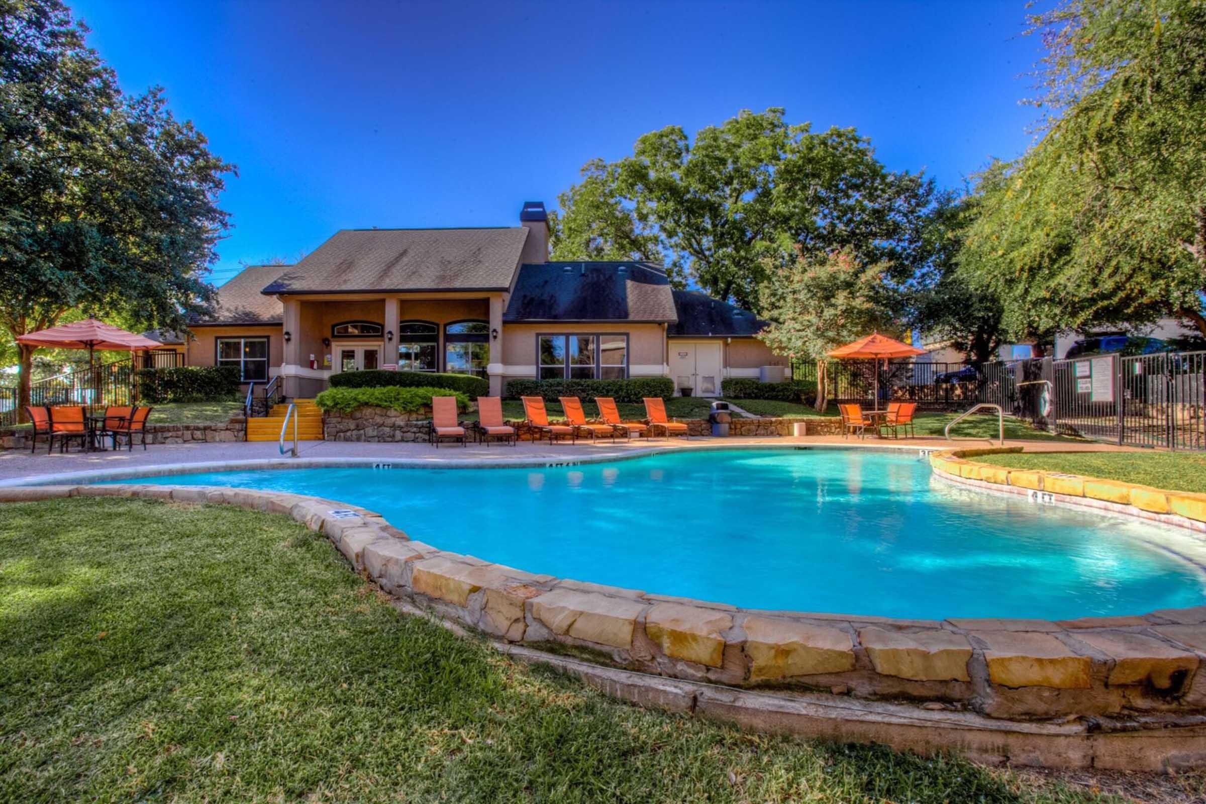 A clear blue swimming pool surrounded by green grass and orange lounge chairs. In the background, a cozy building with large windows and a sloping roof is visible, along with several trees providing shade. Orange umbrellas are set up near the seating area, creating a welcoming outdoor atmosphere.