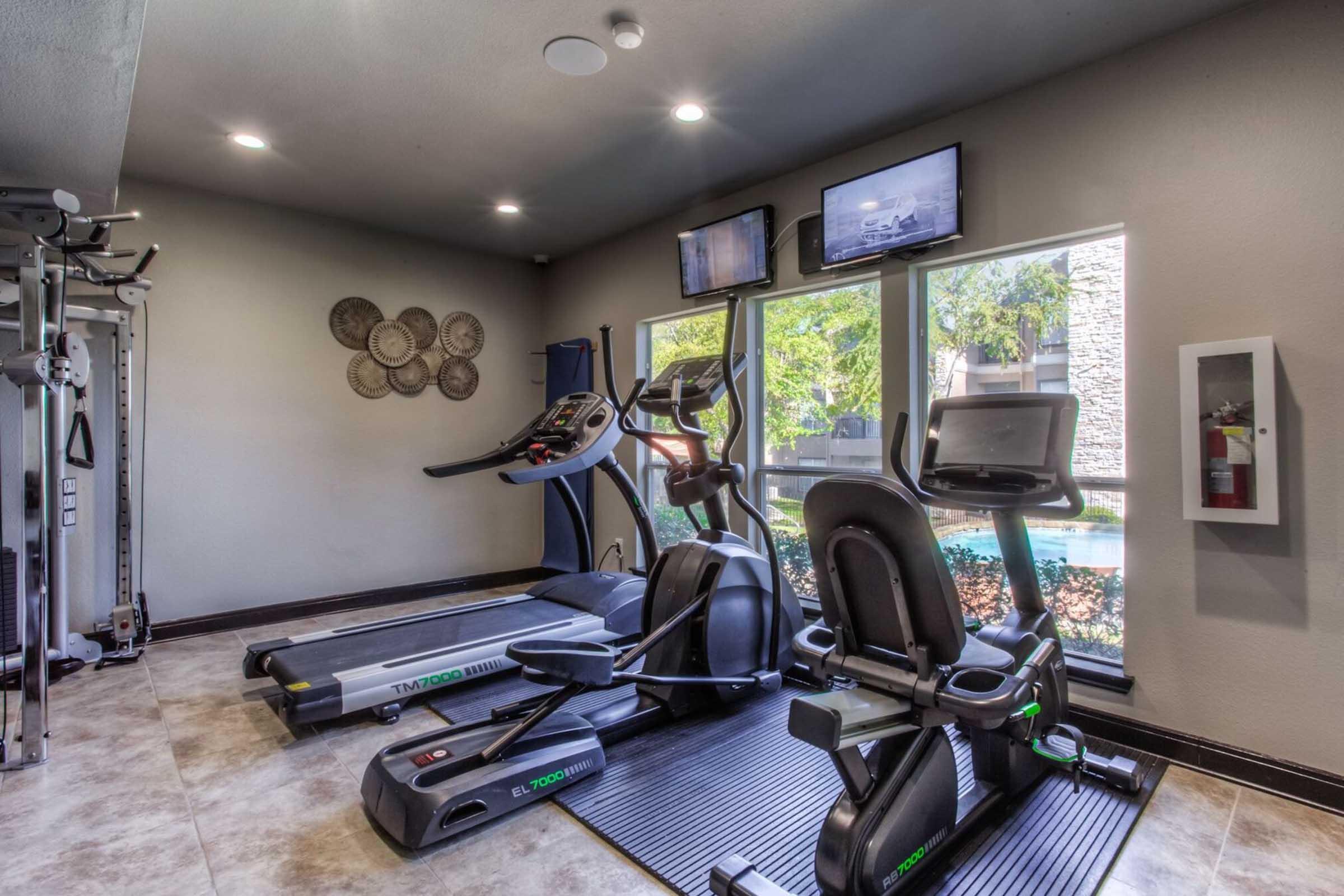A modern gym space featuring two treadmills and an elliptical machine, positioned near large windows that provide natural light. The room is equipped with two flat-screen TVs mounted on the wall and has a sleek, contemporary design with gray walls and tiled flooring.