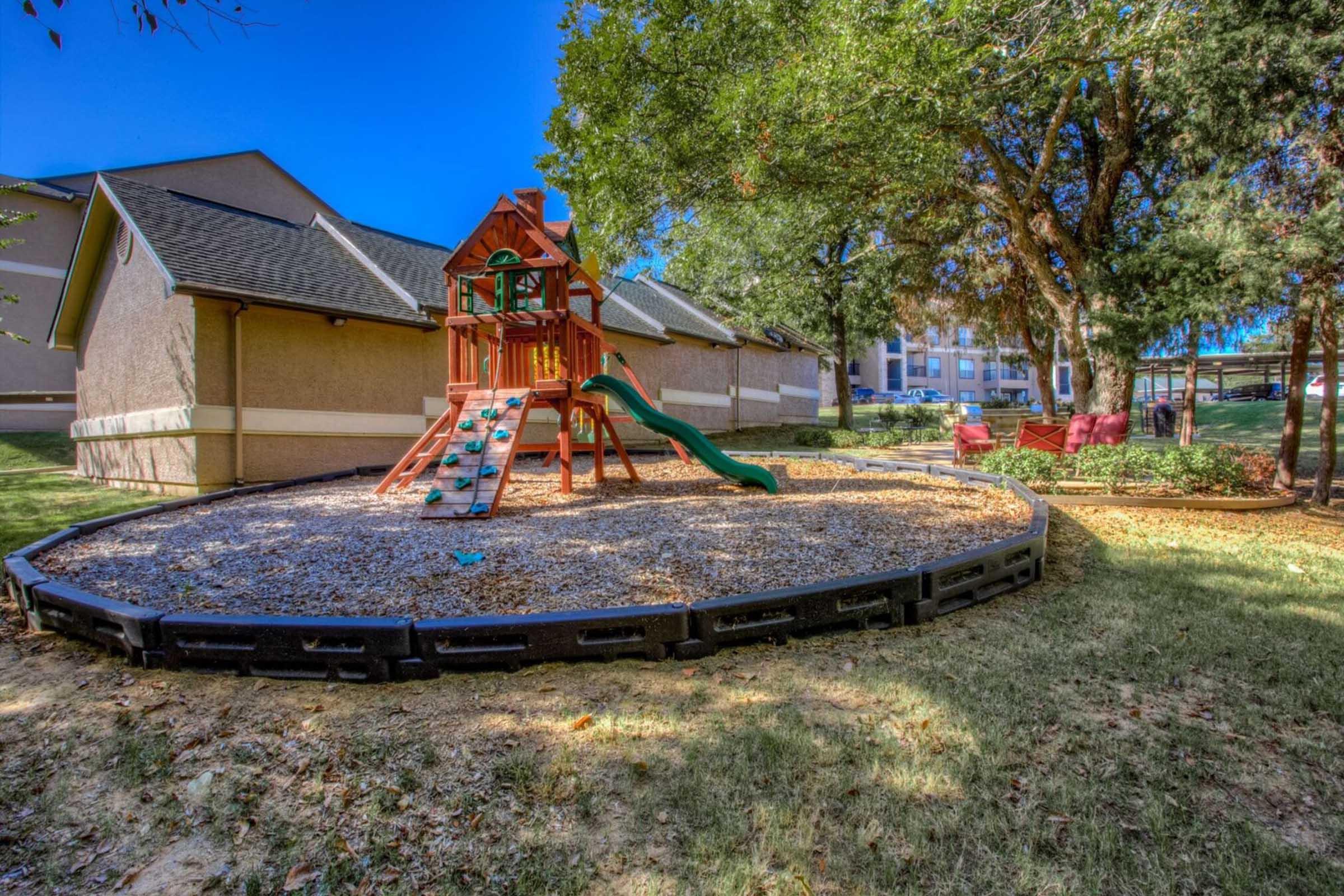 A playground featuring a wooden playset with a slide and climbing structure, surrounded by a gravel surface and grass. In the background, there are buildings and trees providing shade, with a play area visible to the right, creating a safe and inviting outdoor space for children.