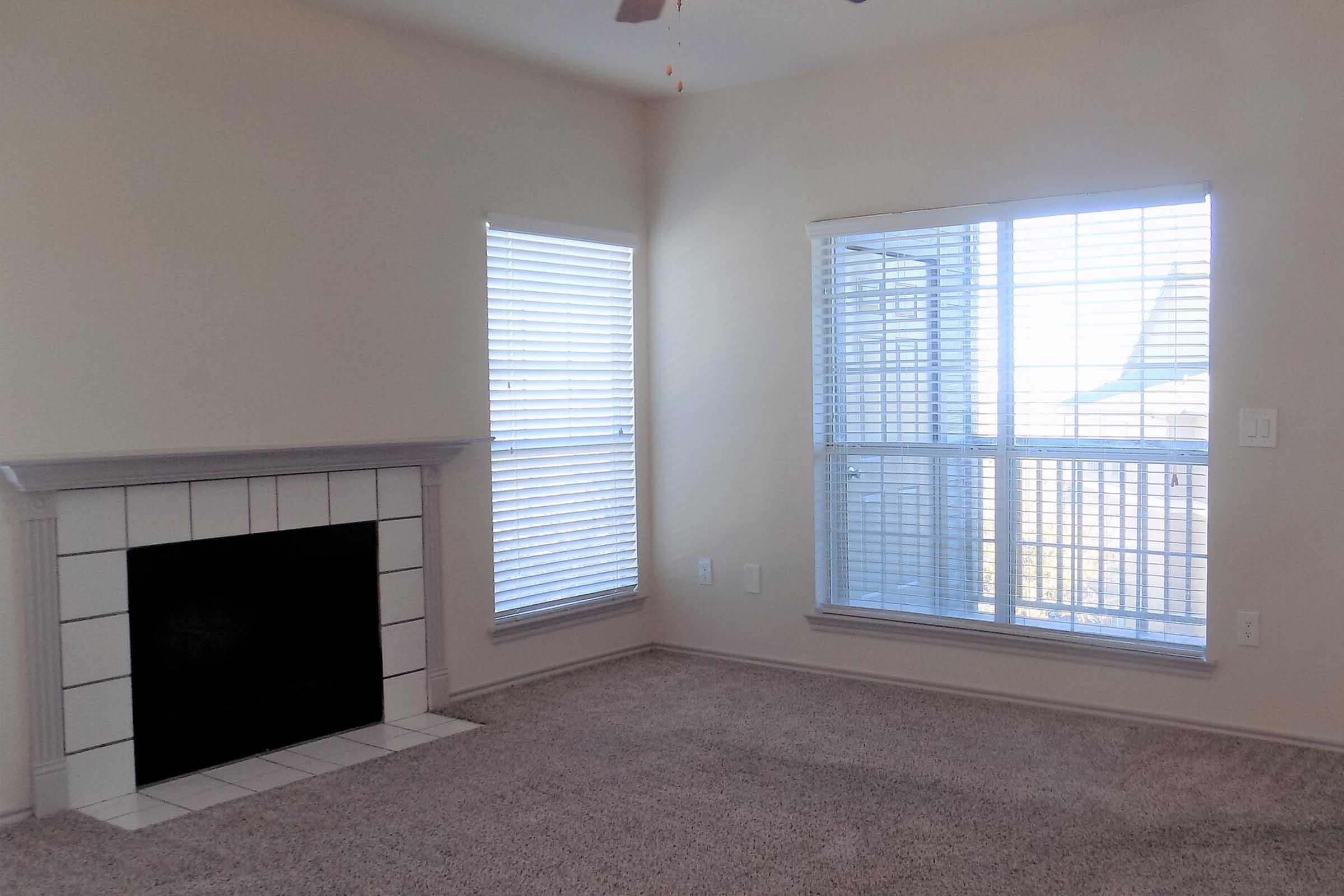 A cozy living room featuring a white tiled fireplace, light beige walls, and large windows with blinds allowing natural light. The carpeted floor enhances the warm ambiance, while the room is bright and inviting, perfect for relaxation or social gatherings.
