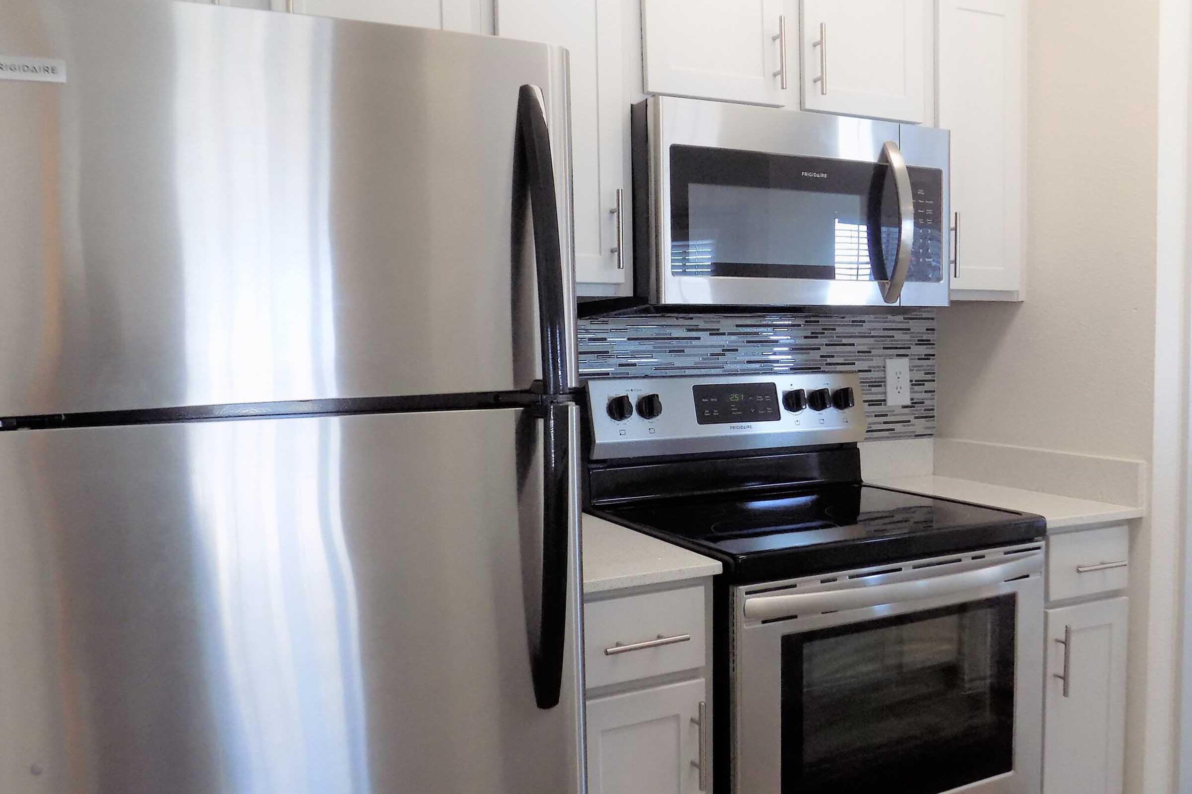 Interior view of a modern kitchen featuring stainless steel appliances including a refrigerator, an oven, and a microwave. The cabinets are white, and a decorative backsplash with a mosaic design adds style. The countertop is light-colored, complementing the overall contemporary design.