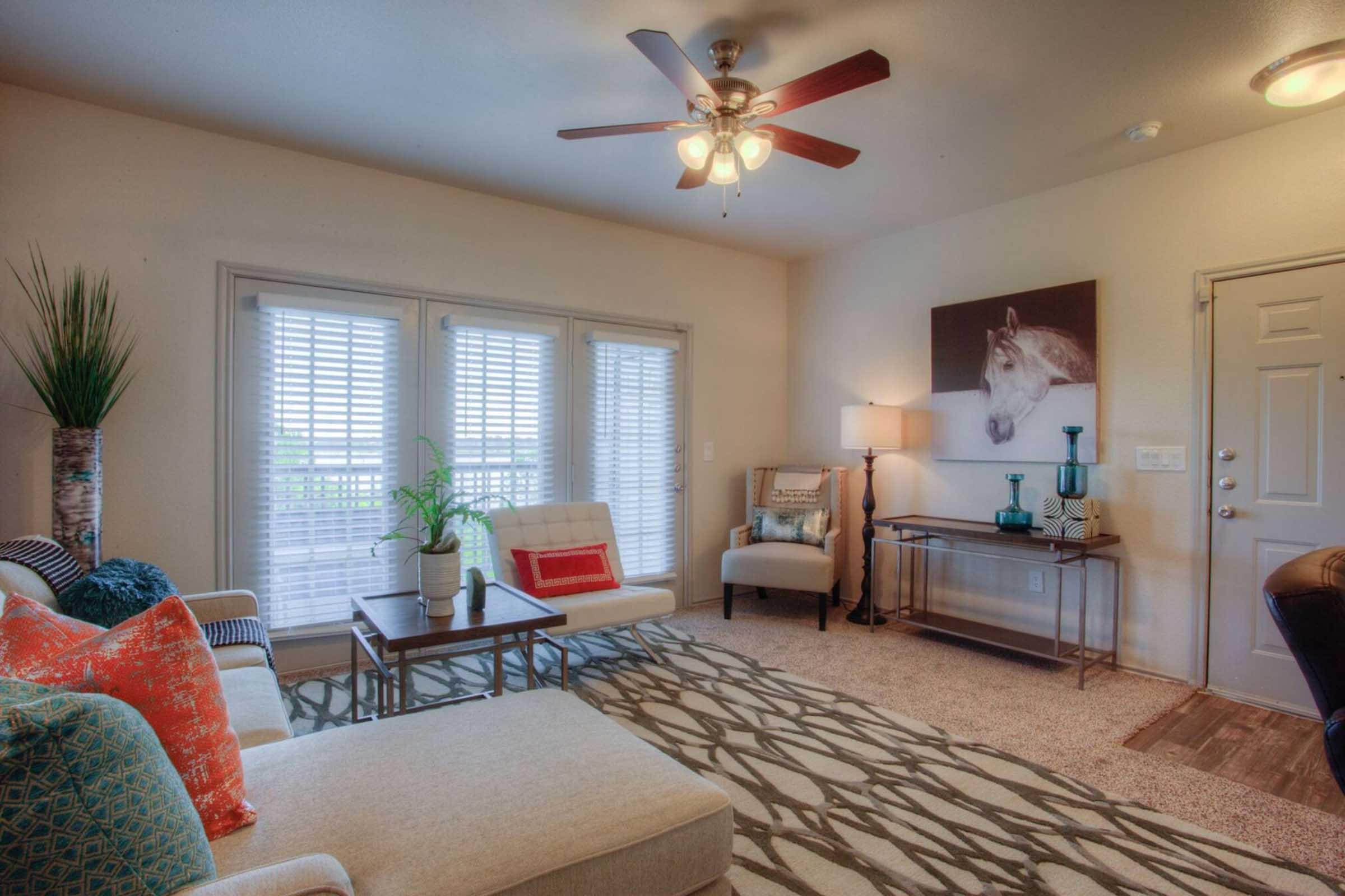 A well-decorated living room featuring a light-colored sofa with colorful throw pillows, a wooden coffee table, and a chair beside it. There is a decorative plant, a ceiling fan, and large windows with white shutters letting in natural light. A painting of a horse adorns the wall.