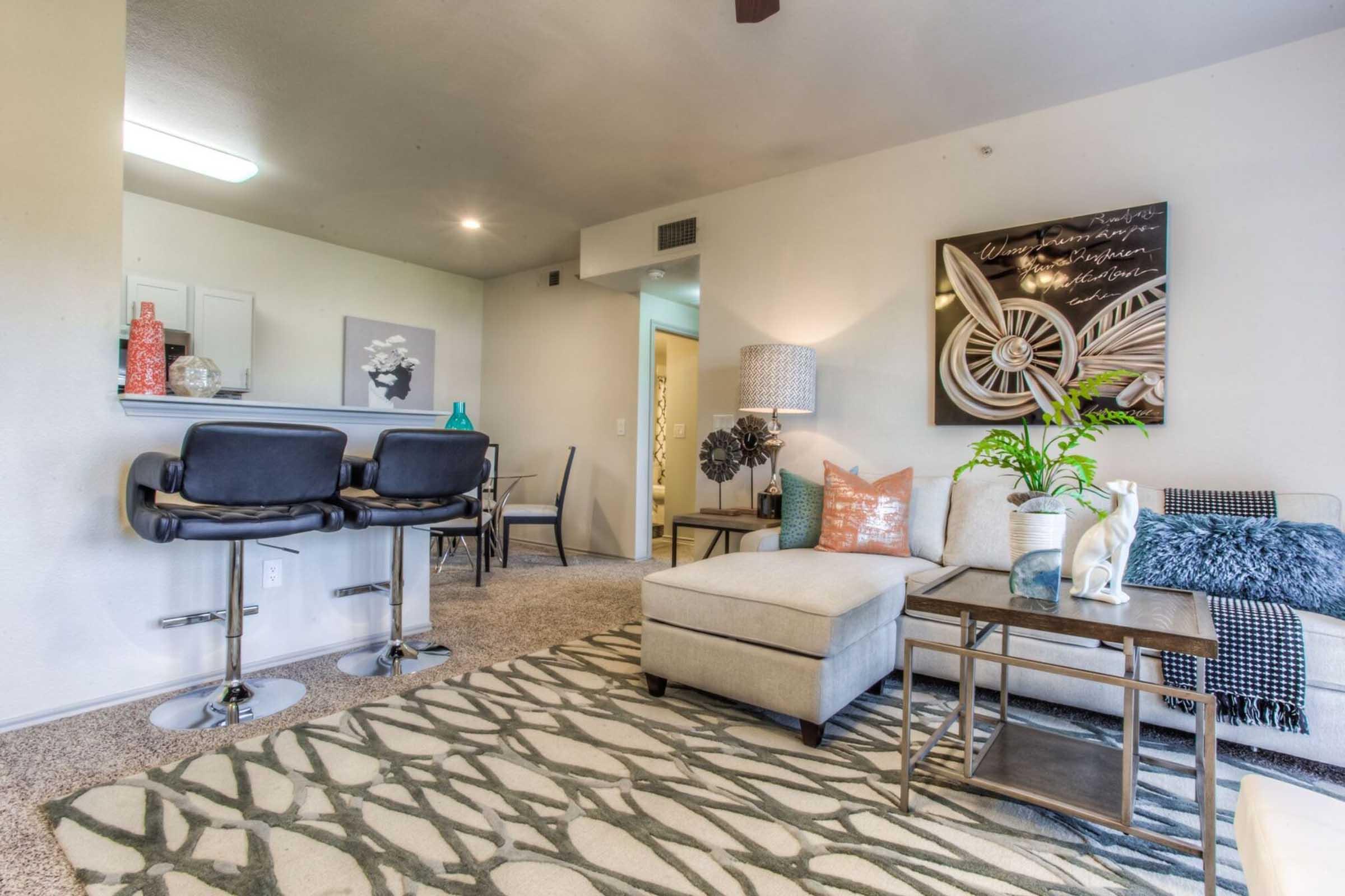A modern living room featuring a light-colored sectional sofa, a decorative rug with a leaf pattern, and a coffee table. There are stylish accents like a lamp and decorative pillows. A bar area with two black stools is visible, along with a wall art piece and indoor plants, creating a cozy and inviting atmosphere.
