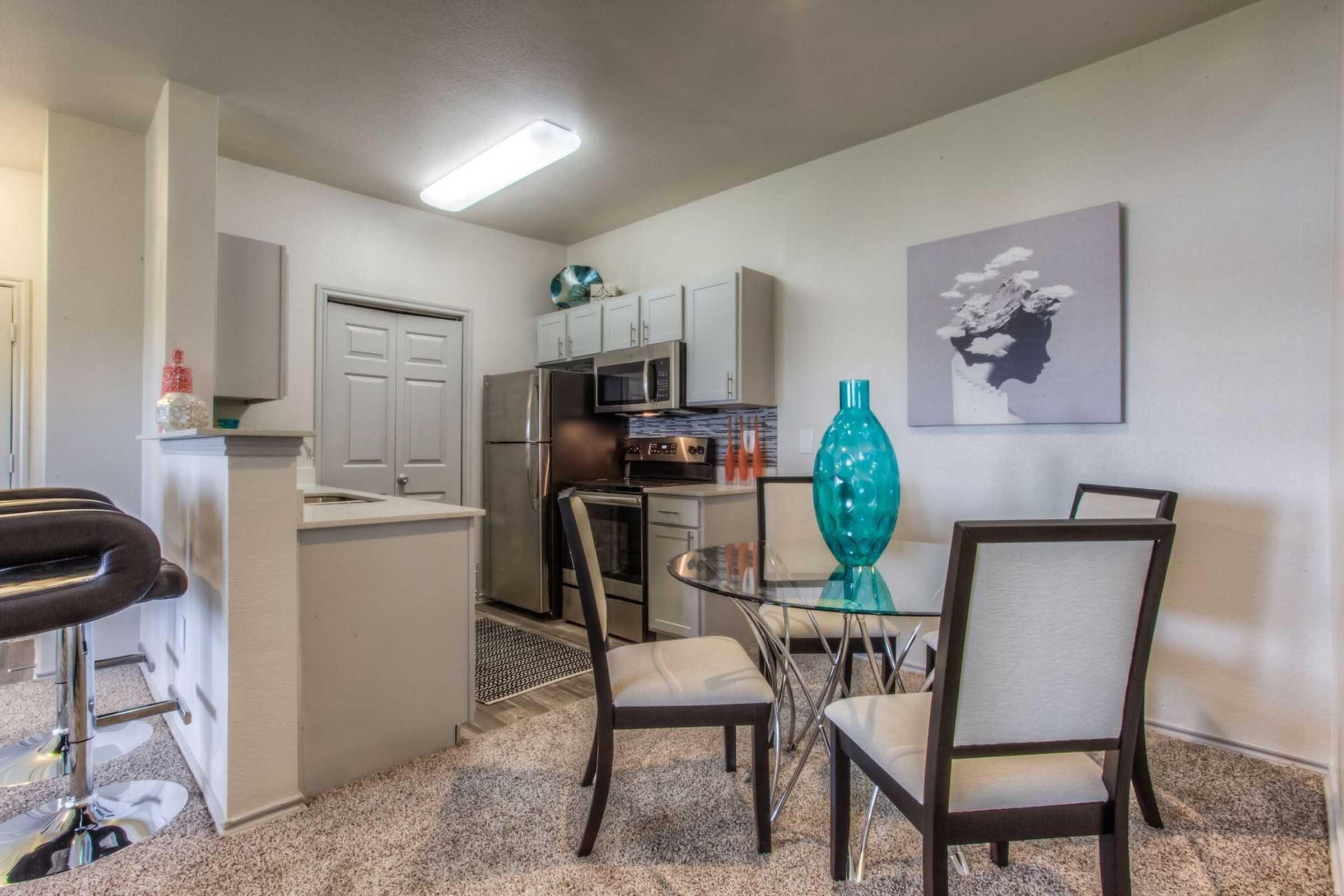 A modern kitchen and dining area featuring a round glass table with four chairs, a teal decorative vase, stainless steel appliances, and light gray cabinets. The walls are painted in neutral tones, and there is a piece of artwork on the wall. The floor is covered with plush carpeting.
