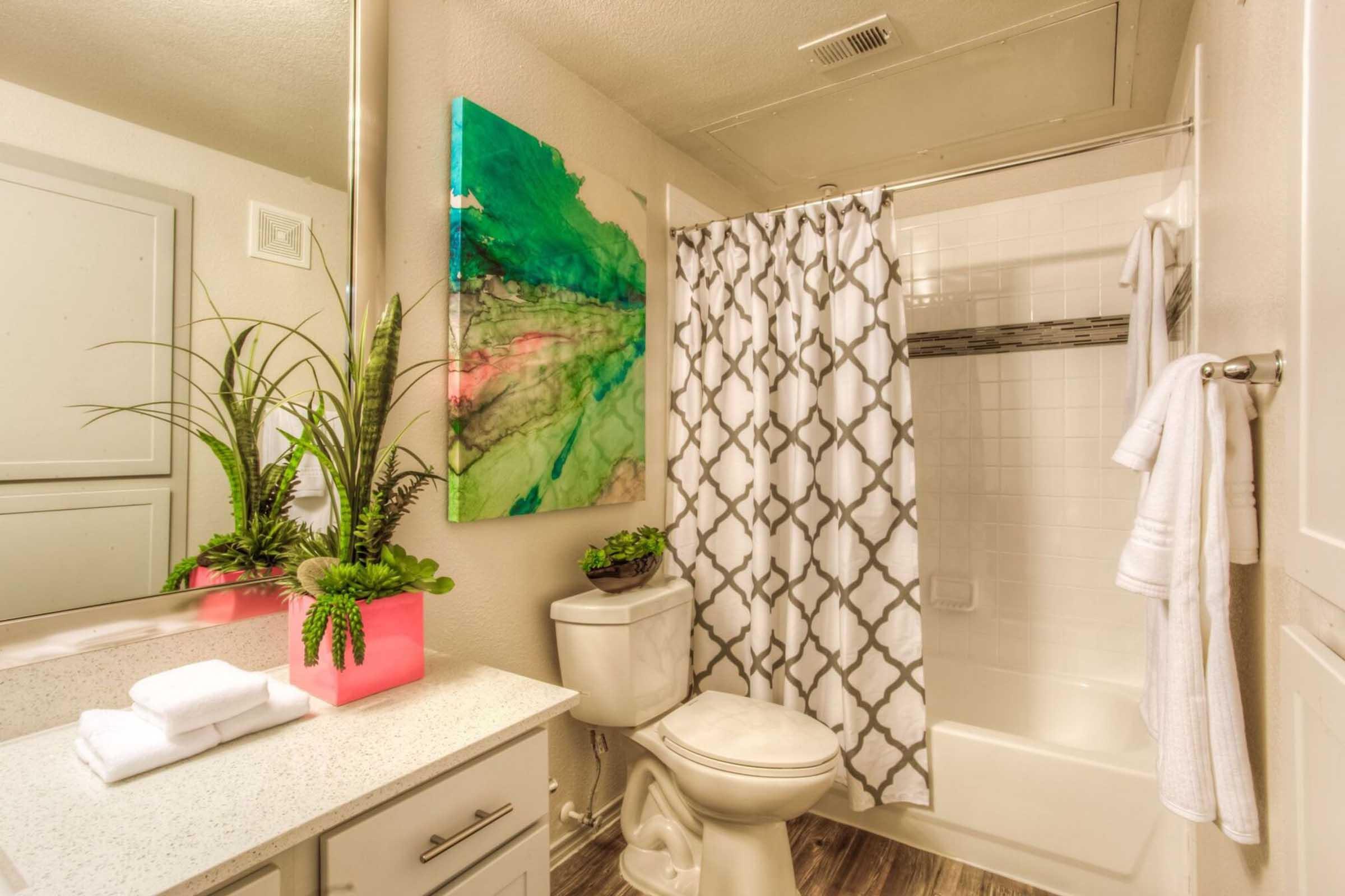 A modern bathroom featuring a white bathtub with a shower curtain, a toilet, and a sink with a stylish countertop. Decorative elements include potted plants and neatly arranged towels, along with a colorful abstract painting on the wall that adds a pop of color to the space.