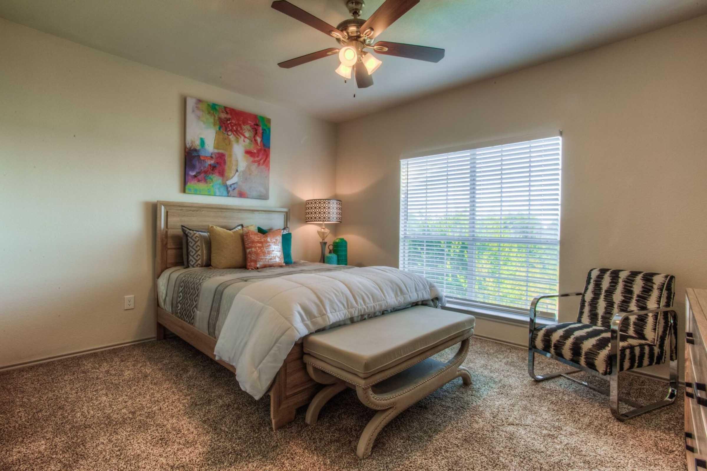 A cozy bedroom featuring a queen-sized bed with decorative pillows, a stylish bench at the foot, and a contemporary chair. A large window with blinds offers natural light, while a colorful piece of art adorns the wall. The floor is covered in plush carpeting, creating a warm atmosphere.