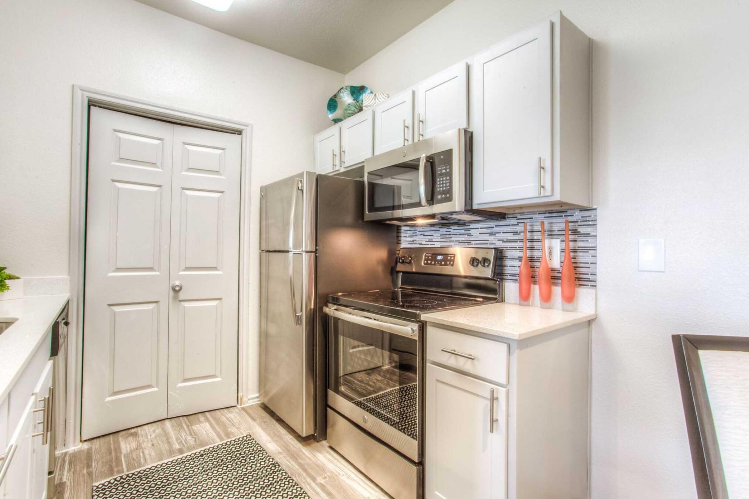 Modern kitchen featuring stainless steel appliances, including a refrigerator and oven. White cabinetry and a light backsplash complement the space. A door leads to another area, while decorative vases in vibrant colors add a touch of style. The floor is finished with wood-like tiles.