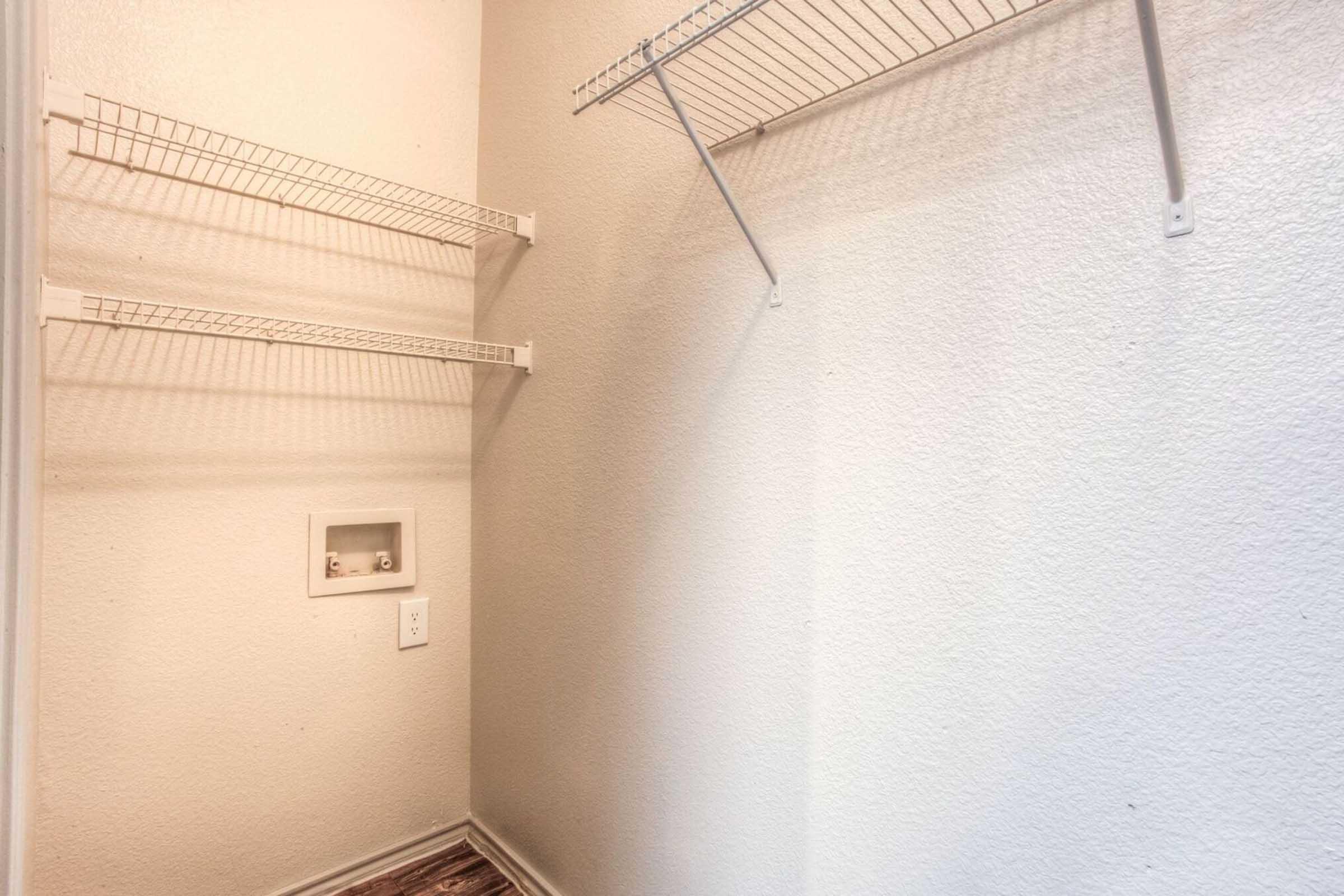 Empty closet with light gray walls, featuring two white wire shelves on the left and a single wire shelf on the right. A small electrical outlet is visible near the floor, and the floor appears to be dark-colored wood. The overall space is uncluttered and minimalistic.