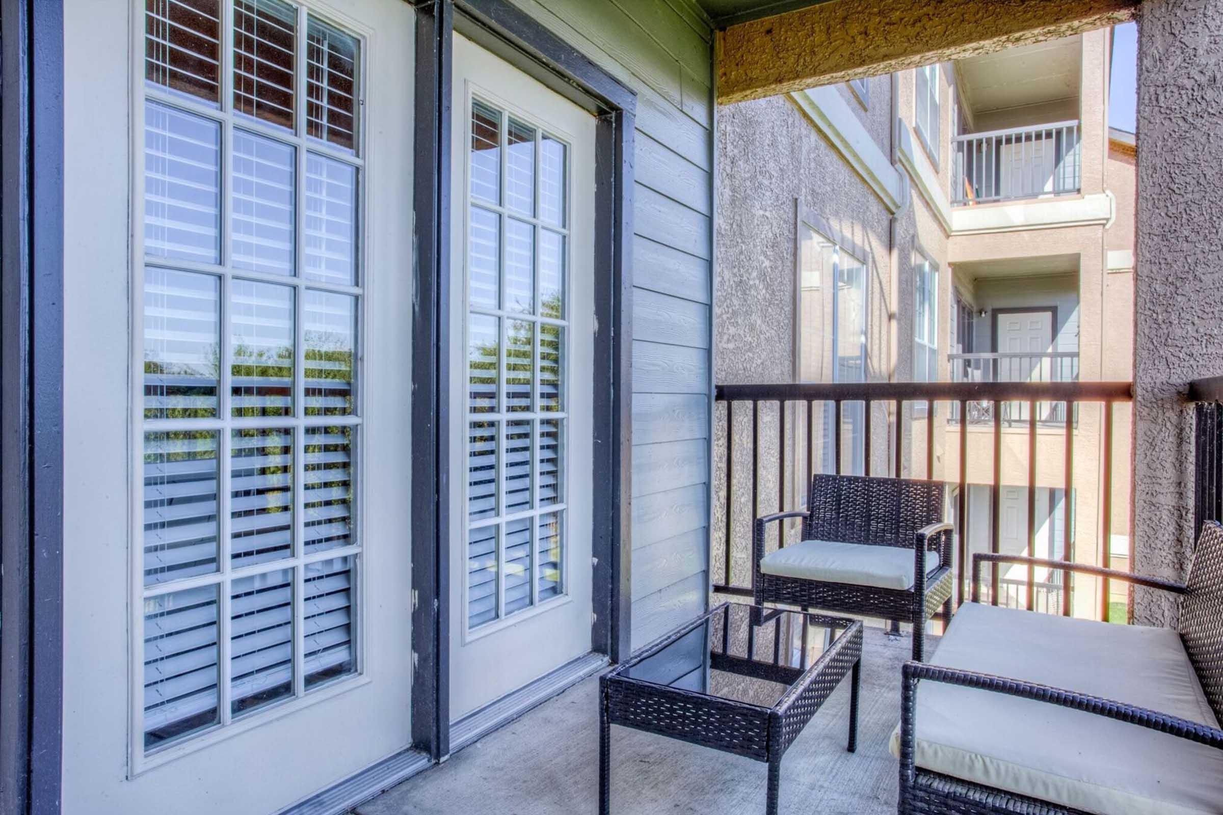 A small balcony featuring two wicker chairs and a glass table, with French doors leading to an interior space. The balcony has a view of other apartment buildings and greenery outside. Natural light illuminates the space, creating a cozy atmosphere.