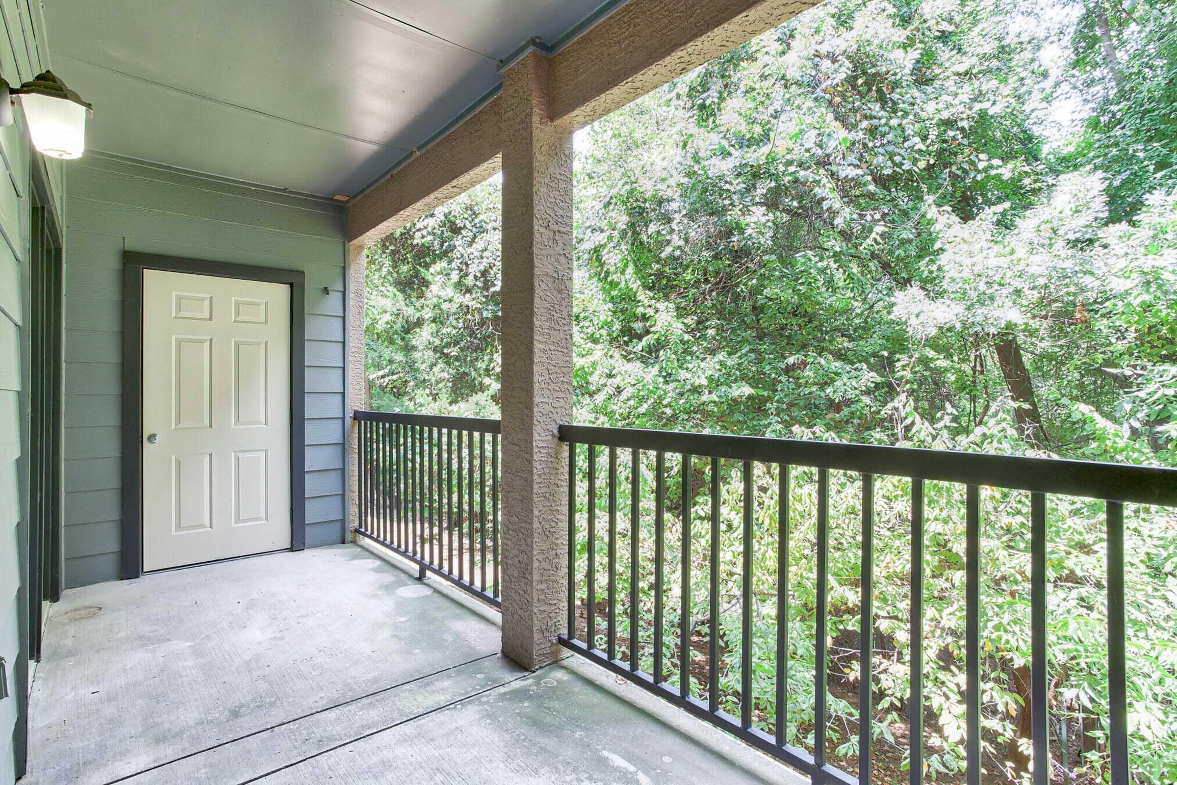 A view of a covered balcony or porch with a railing, featuring gray siding and a white door. The space is surrounded by lush greenery, creating a serene and natural atmosphere. Bright natural light filters in, highlighting the outdoor setting.