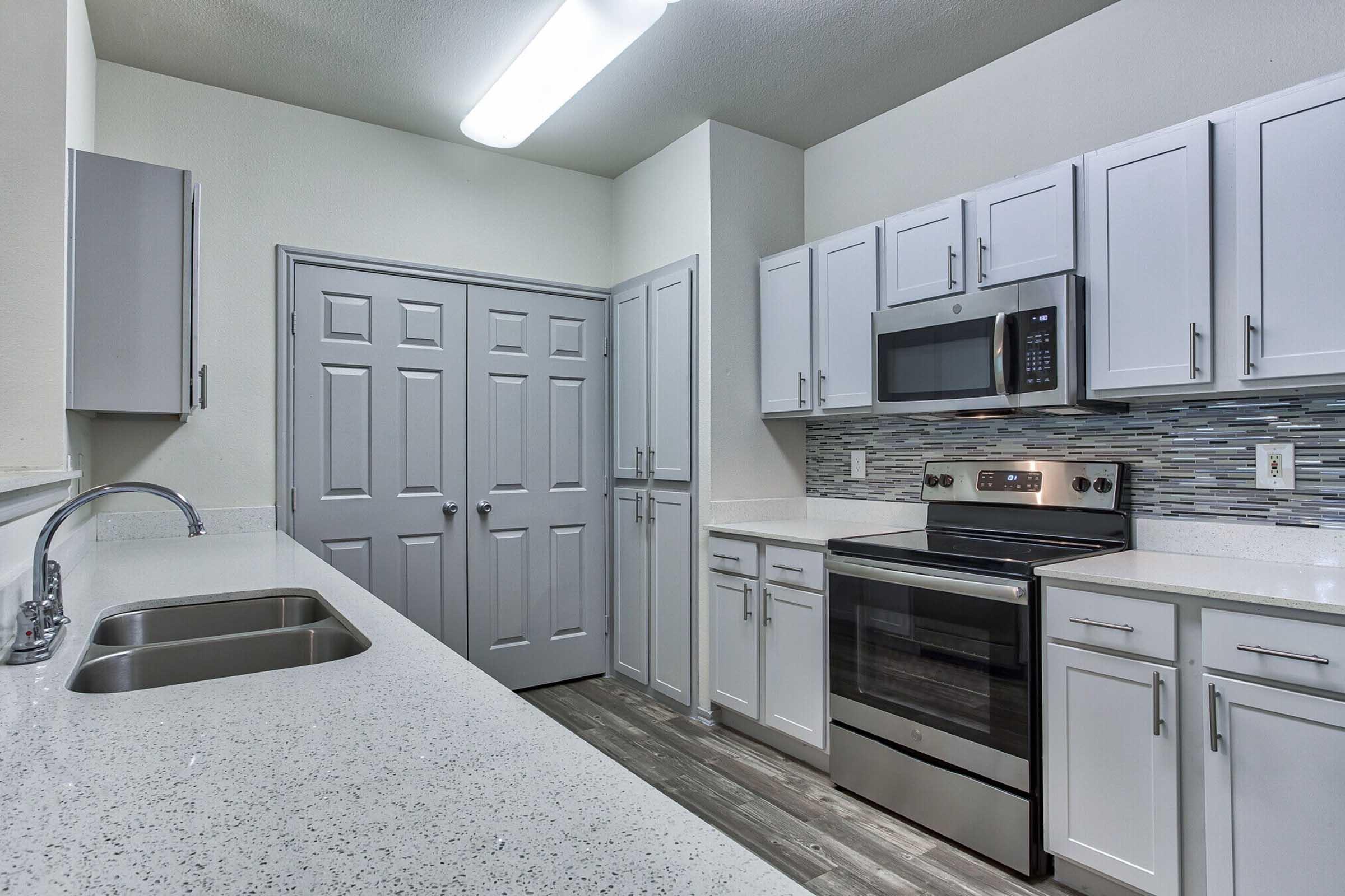 Modern kitchen featuring white cabinetry, a stainless steel oven and microwave, and a double sink with a spacious countertop. The room has a light-colored backsplash and gray flooring, with a door leading to another area. Brightly lit with overhead lighting.
