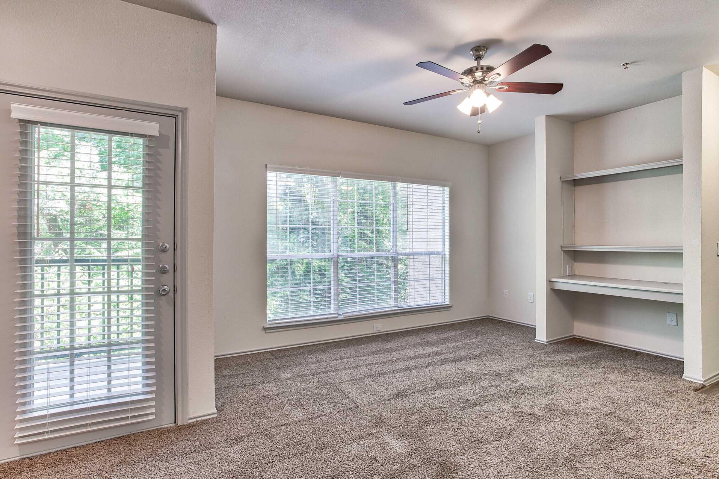 Spacious living room with beige walls and carpeted flooring, featuring a ceiling fan, large window with white blinds allowing natural light, and an adjacent sliding glass door leading to a balcony. A built-in shelf unit is visible on the right side.