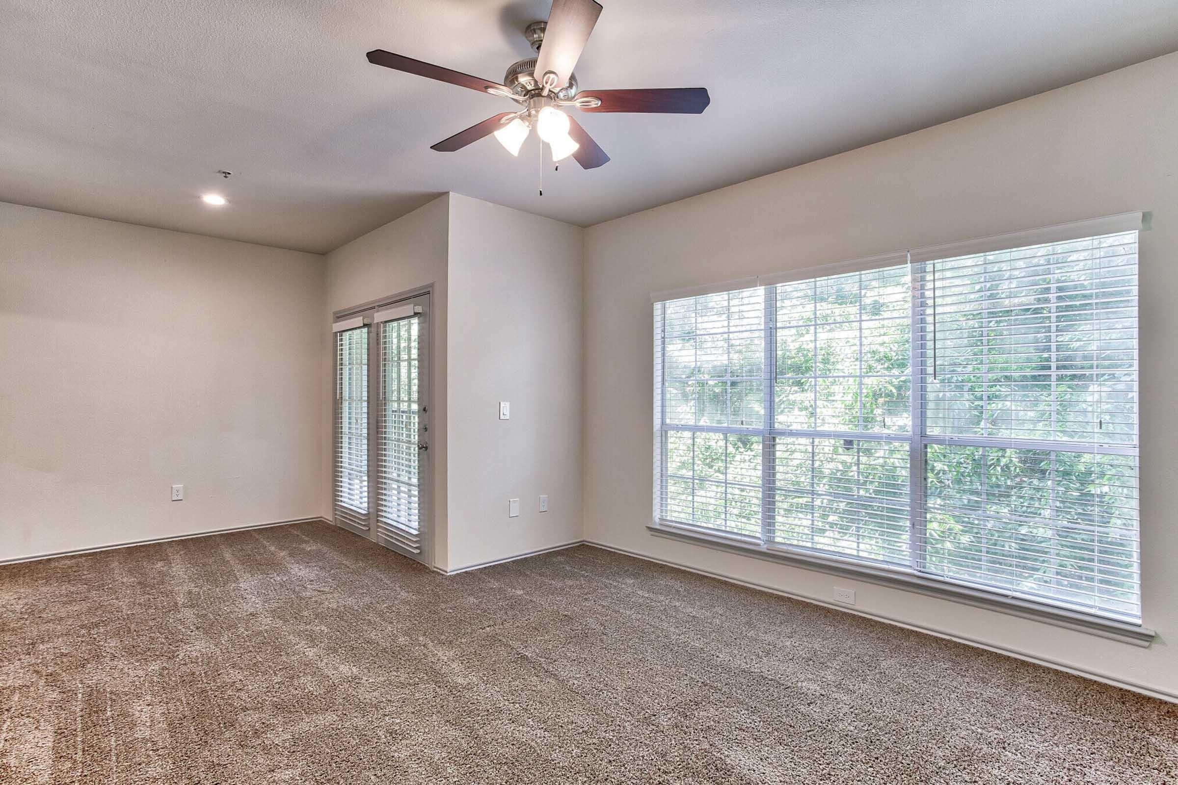 A spacious, empty room with light brown carpet, a ceiling fan with light, and large windows with white blinds letting in natural light. Double doors lead to an outdoor space, and the walls are painted a light neutral color, creating a bright and inviting atmosphere.