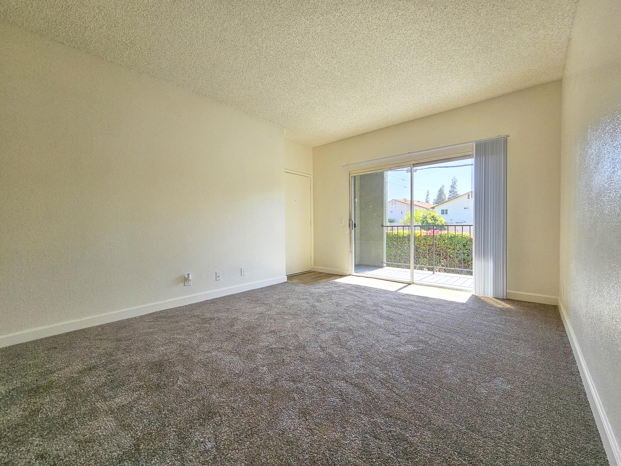 A bright, empty living room with beige carpet and light-colored walls. A large sliding glass door leads to a small balcony, allowing natural light to fill the space. The room appears spacious and clean, with no furniture or decorations present.