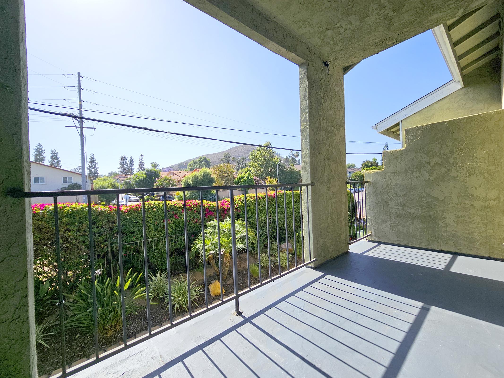 View from a balcony featuring a railing, with a backdrop of green bushes and trees. In the distance, there are hills under a clear blue sky. The sunlight casts shadows on the ground, creating a peaceful outdoor atmosphere.