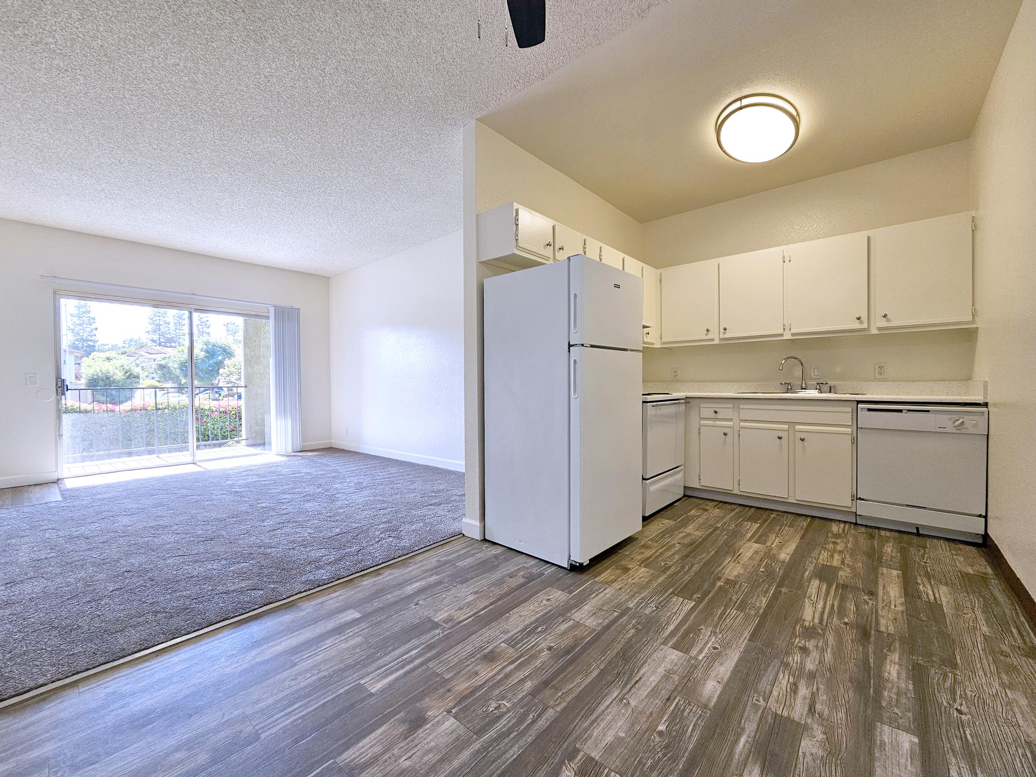 A spacious apartment interior featuring a kitchen with white cabinets, a refrigerator, and a dishwasher. The floor has wooden laminate, with carpeted living area visible in the foreground. A sliding glass door leads to a balcony with views of greenery outside. Bright, open layout enhances the sense of space.