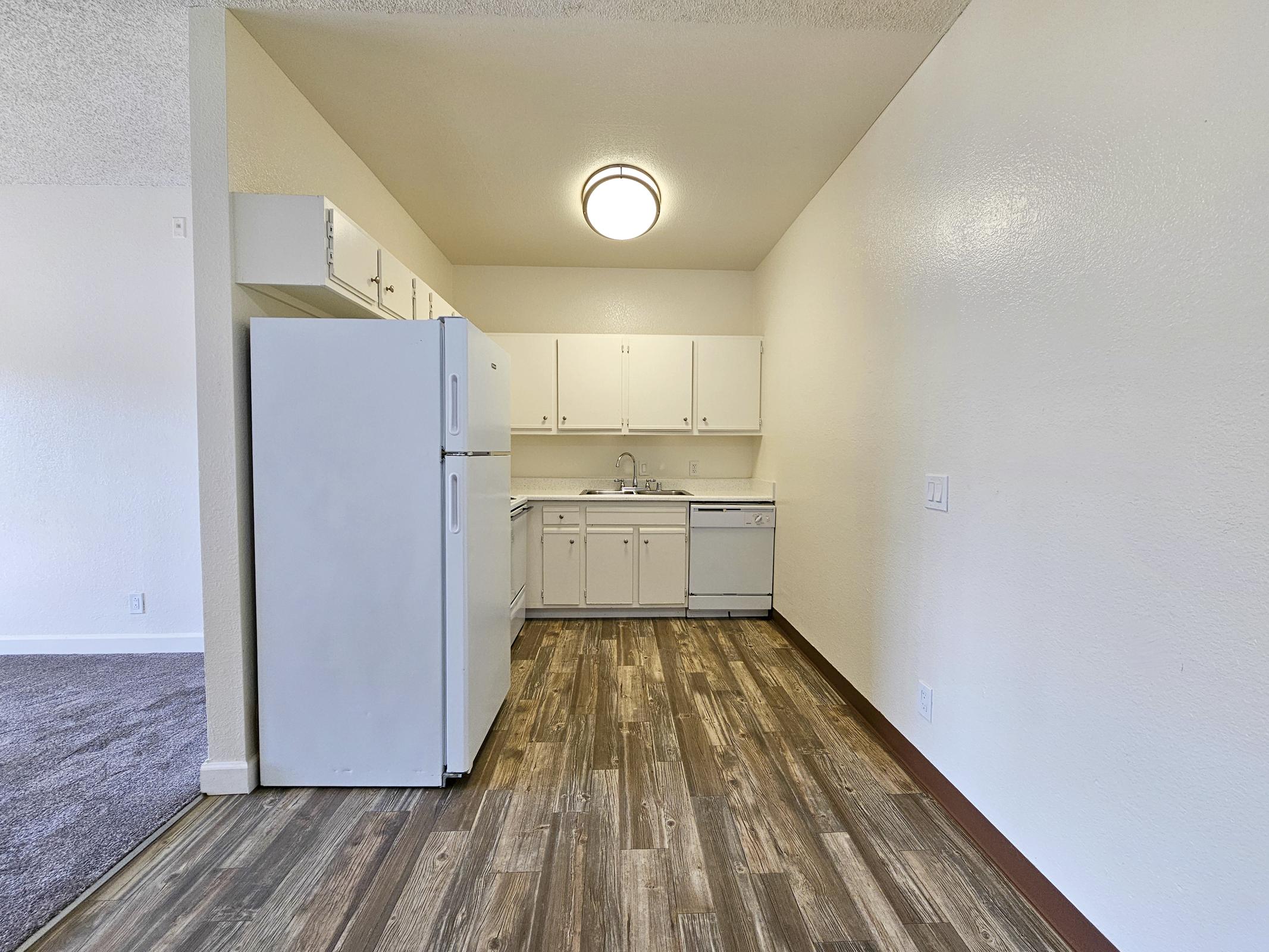 A modern kitchen with white cabinetry, a large white refrigerator, and a built-in dishwasher. The flooring is a stylish wood laminate, and there's a round ceiling light fixture. The kitchen is well-lit, with walls painted in a light color, creating a spacious and inviting atmosphere.