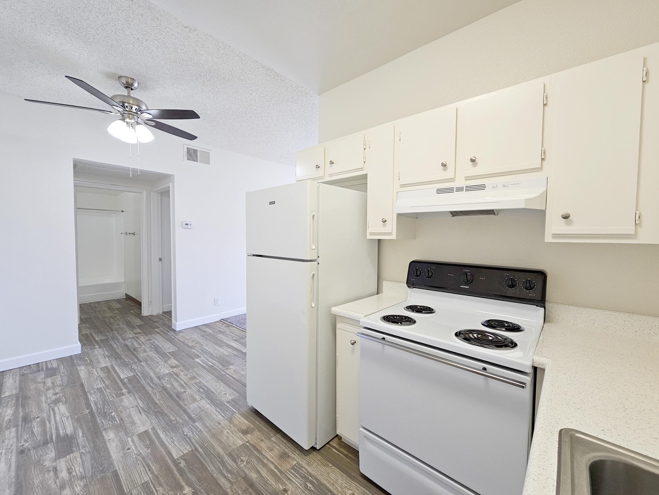 A view of a kitchen space featuring a white refrigerator, a stove with an oven, and white cabinets. The flooring is a light wood-like design, and a ceiling fan is visible. The background shows an open space possibly leading to a living area. The overall ambiance is bright and clean.