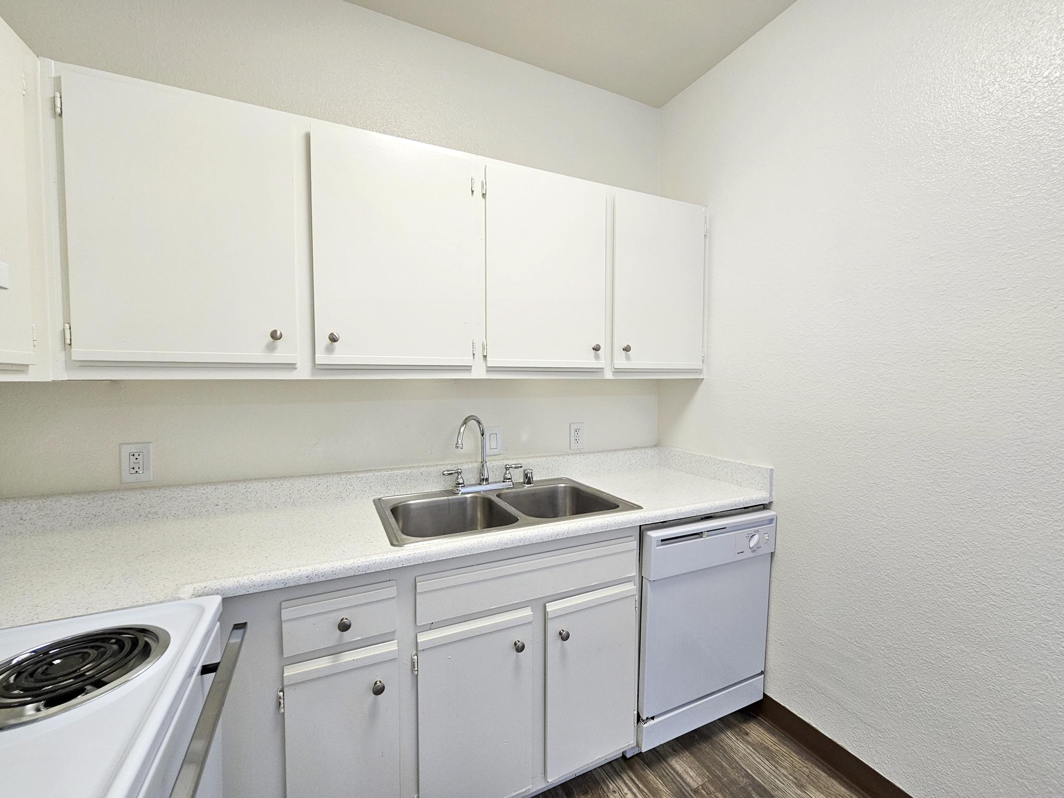 A modern kitchen featuring white cabinets, a dual sink, and a dishwasher. The countertop is light-colored, and there is an electric stove visible in the foreground. The walls are painted in a neutral tone, and the floor has a wood-like finish, creating a clean and tidy cooking space.