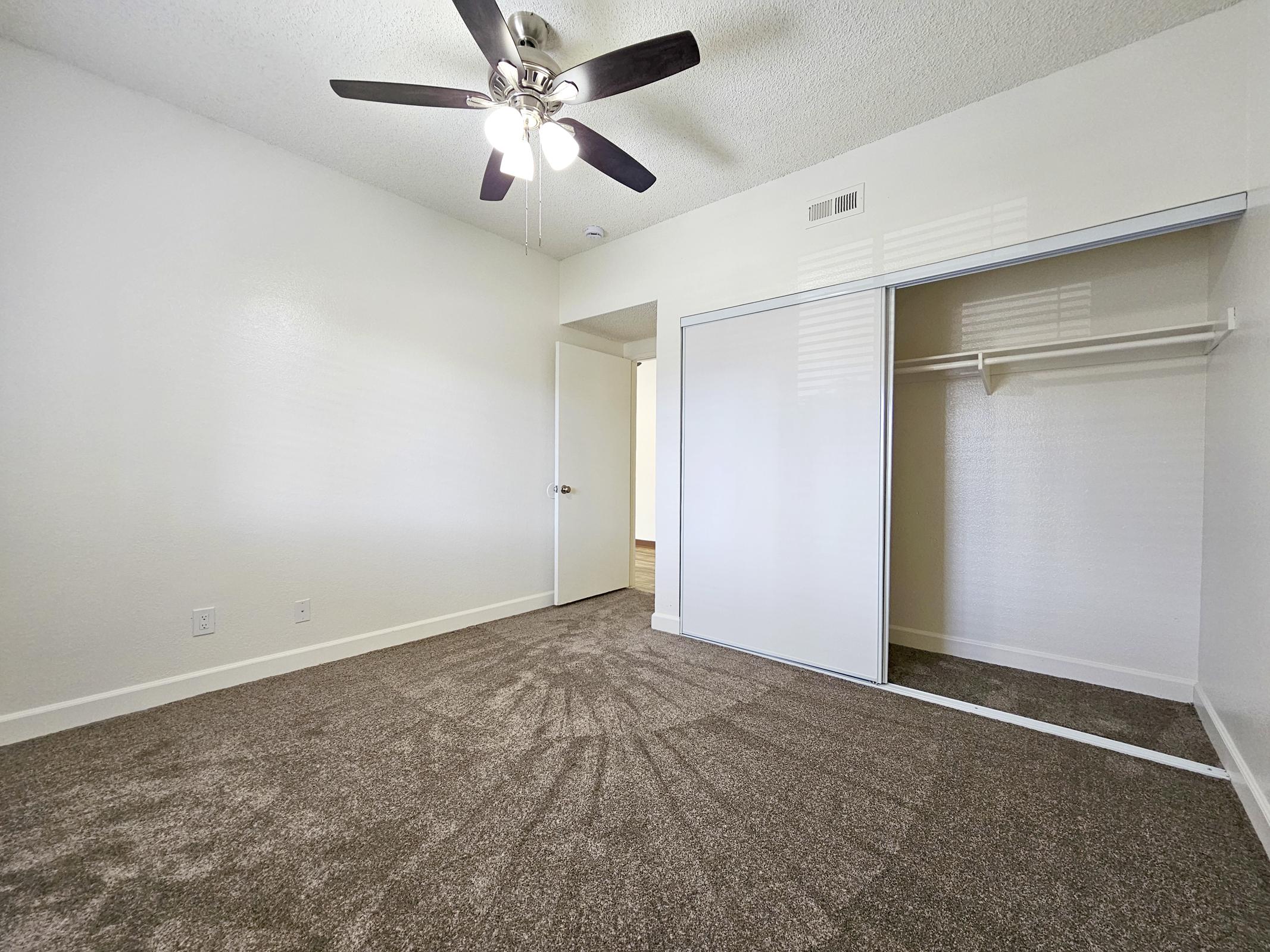 Empty room with beige carpet and white walls. A ceiling fan with three blades hangs from the ceiling. On one side, there's a closet with sliding doors partially open, revealing a hanging rod. A doorway leads to another part of the space, illuminated by natural light.